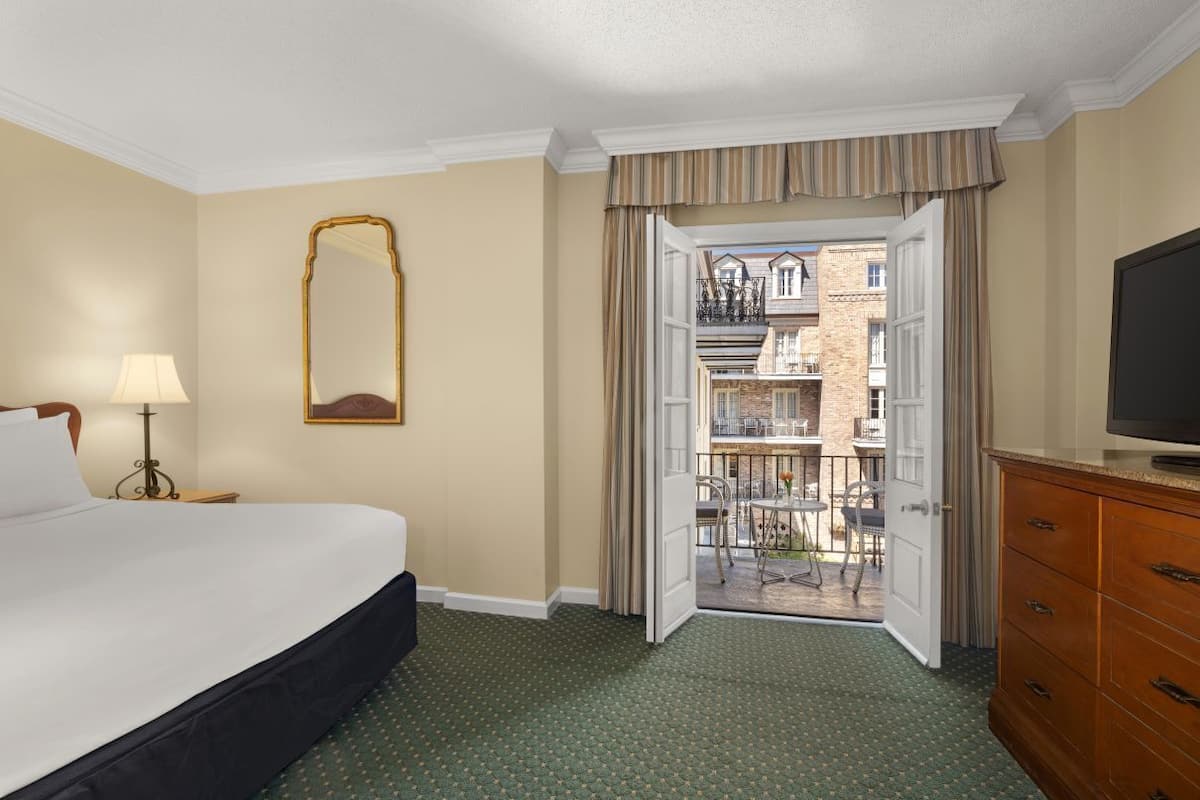 Habitación de hotel con cama, televisión, cómoda, espejo y balcón con mobiliario exterior.