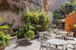 Een terras buiten met rieten meubilair en lichtjes, omringd door weelderige begroeiing en een bakstenen muur.