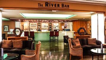 A bar area with a sign reading 'The River Bar,' featuring a bar counter, seating with chairs and a sofa, and a polished wooden floor.