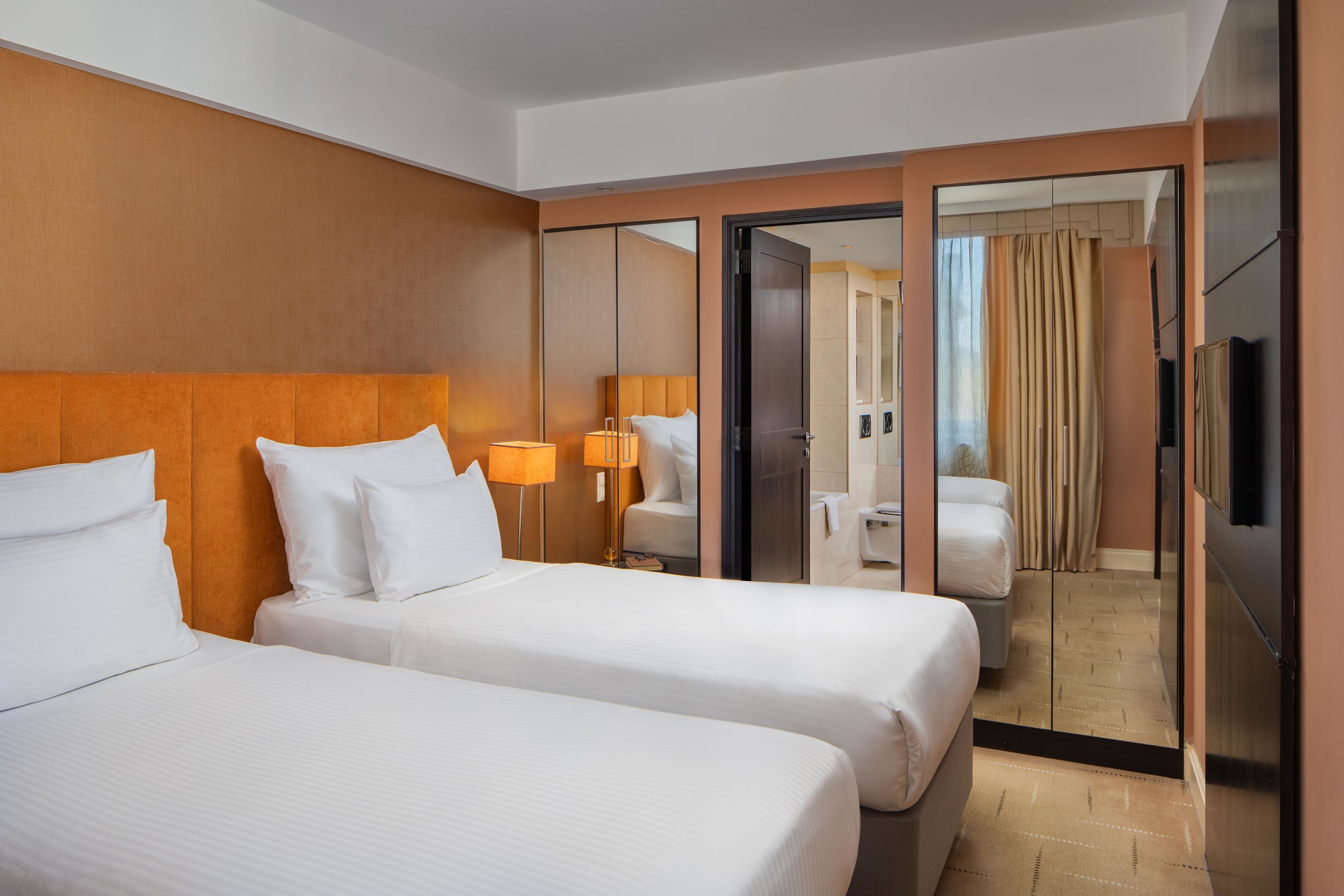 Habitación de hotel con dos camas, un espejo grande, un televisor y un baño visible a través de una puerta corredera.