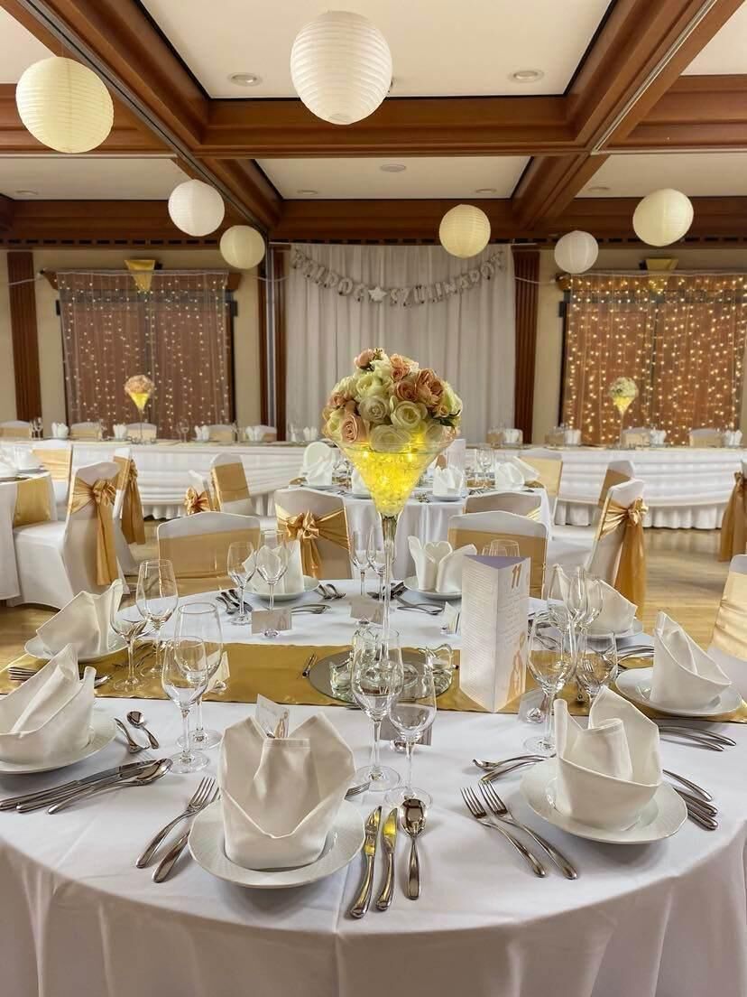 A banquet hall with round tables set for a formal event, featuring white tablecloths, gold sashes, and a central floral arrangement.