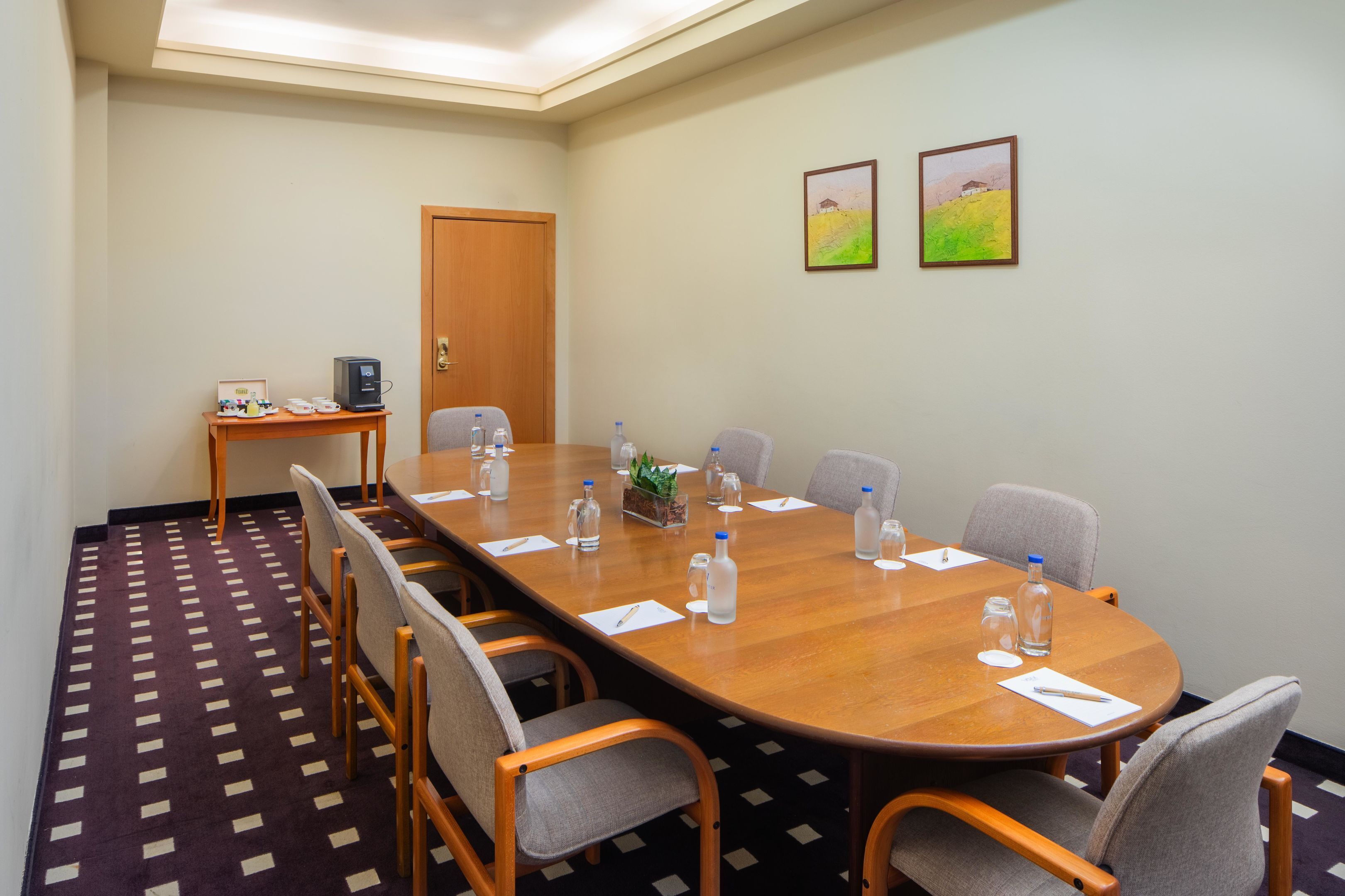 Un salón de conferencias con una mesa larga, sillas, botellas de agua y una pequeña mesa con una computadora y una planta.