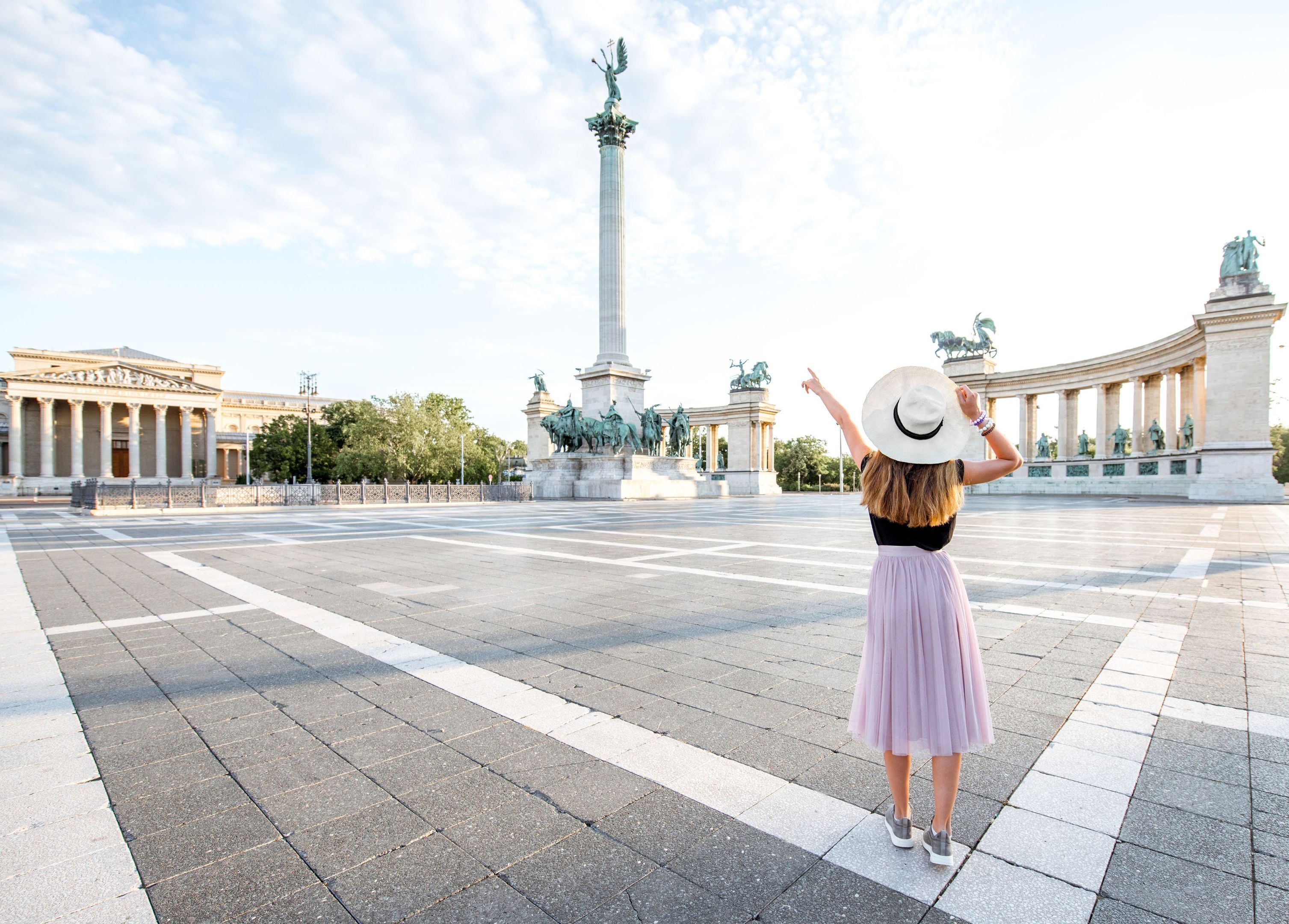 Eine Frau mit Hut und Rock steht auf einem Platz, im Hintergrund ein Denkmal und klassische Architektur.