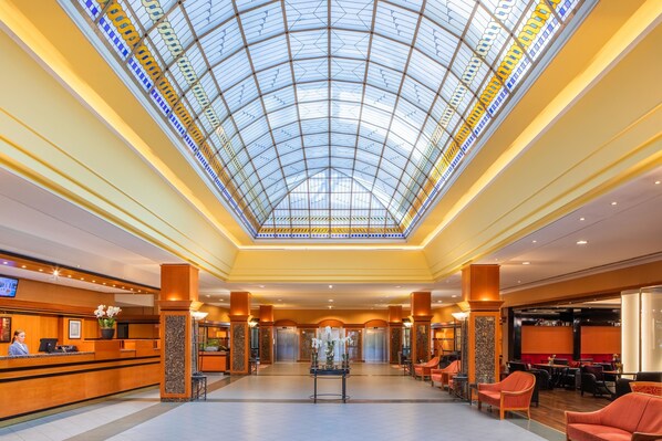 A hotel lobby with a glass ceiling, reception desk, seating area, and elevators.