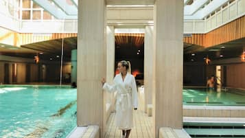 A person in a white robe standing between two pools in a modern indoor setting with large pillars and a tiled floor.