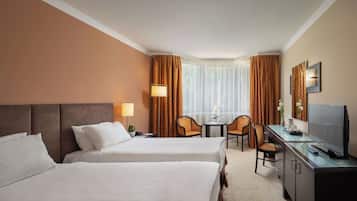 Habitación de hotel con dos camas, un escritorio con televisor, una silla, una mesita de noche con lámpara y una ventana con cortinas.