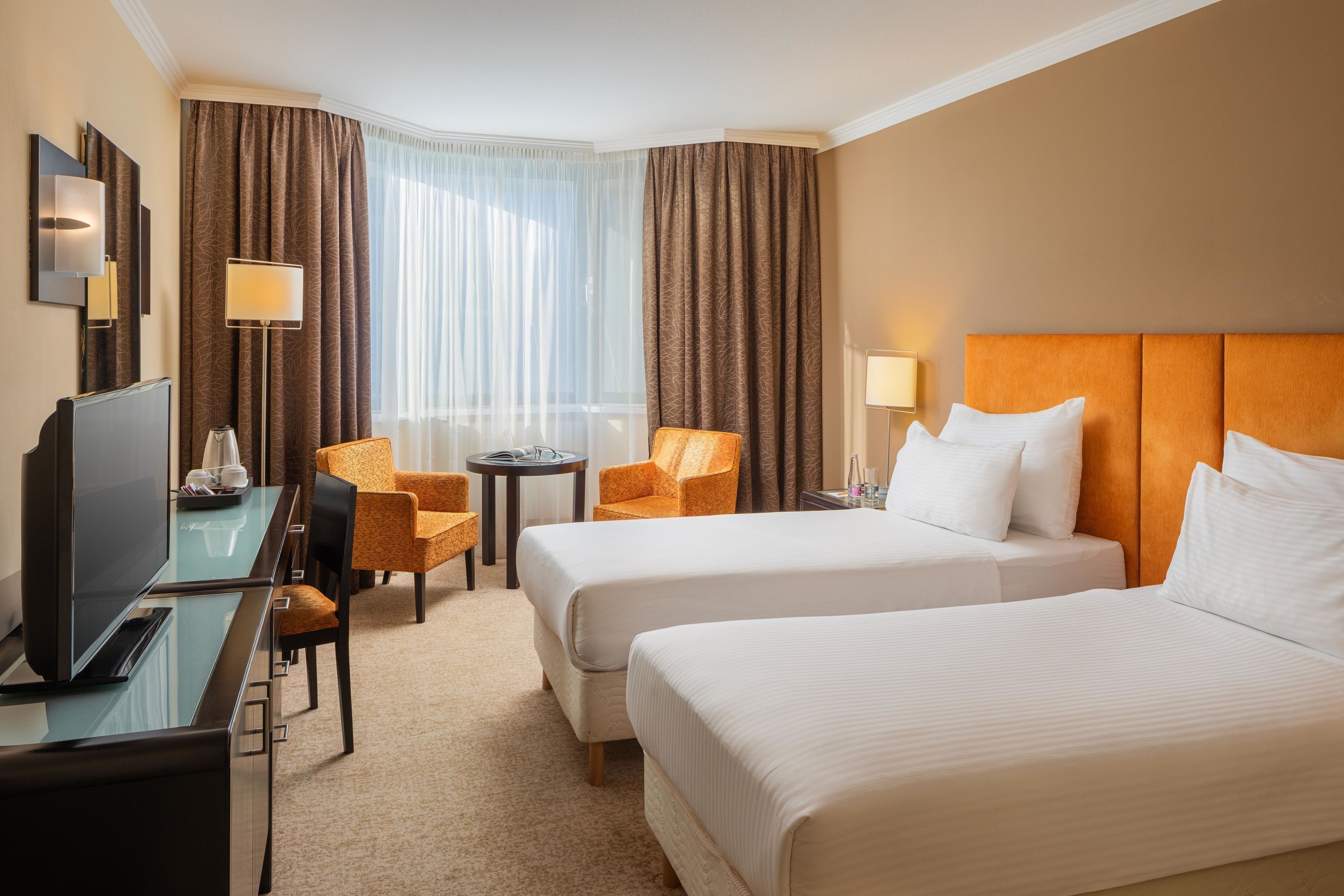 A hotel room with two beds, a desk with a TV, a chair, a small table, and a lamp.