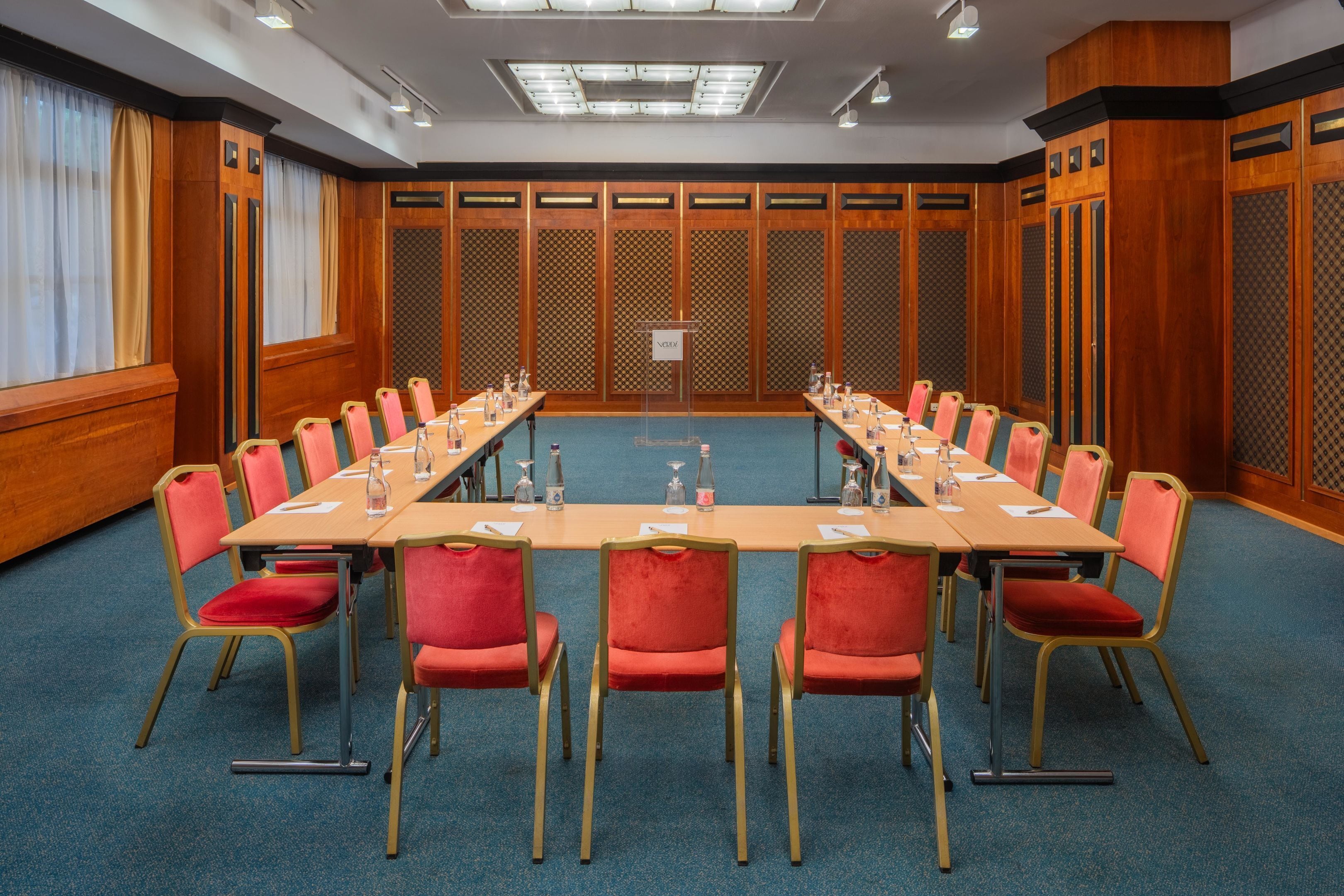Een congreszaal met een lange tafel, rode stoelen en een spreekgestoelte.