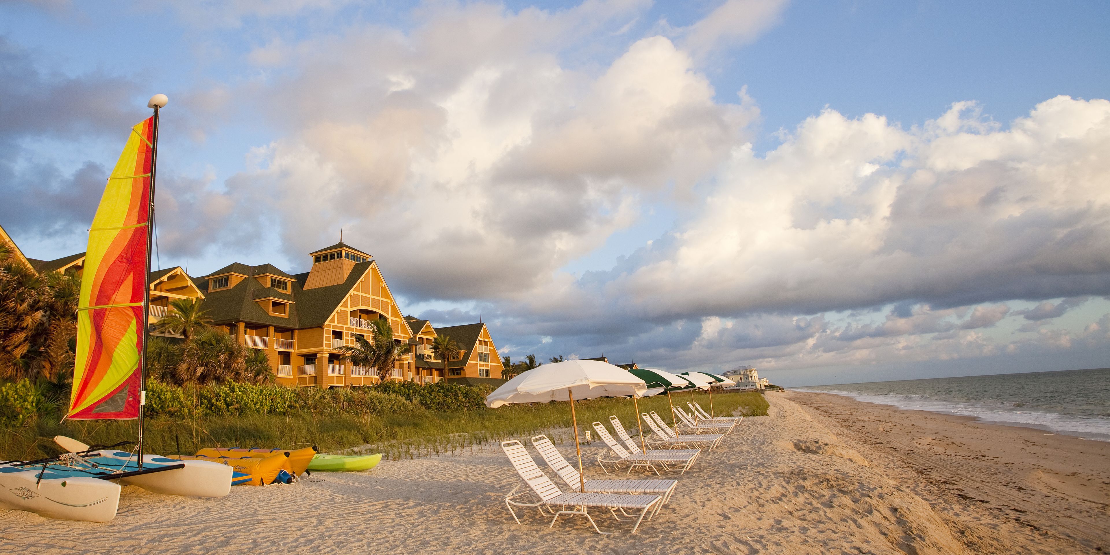 A beach with a row of sun loungers, a sailboat, and a building with a balcony overlooking the ocean.