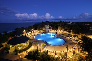Zona de piscina en un resort con un pabellón, sillas de descanso y vista al océano al atardecer.