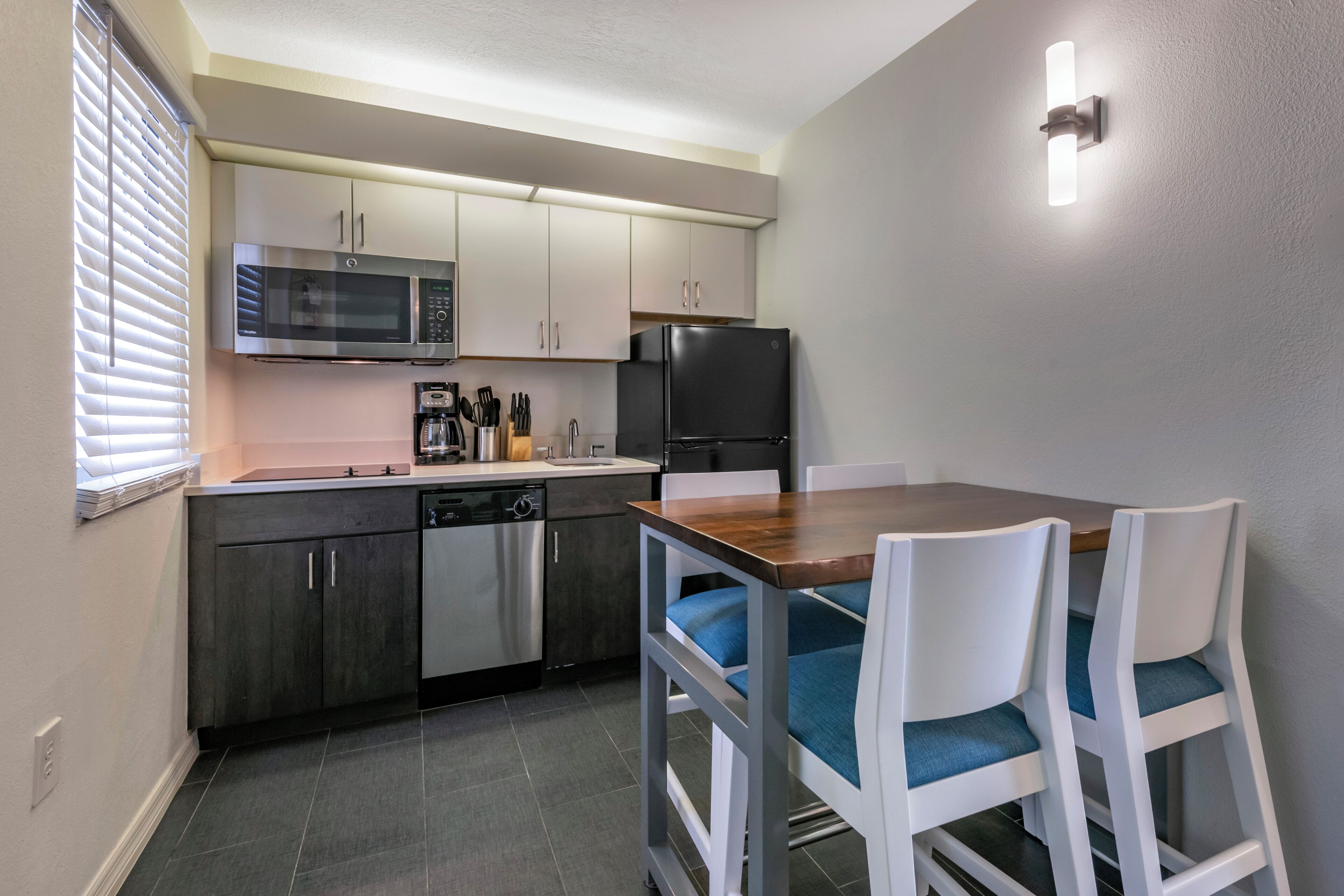 A compact kitchen with a microwave, refrigerator, and dishwasher. There is a dining table with chairs and a window with blinds.