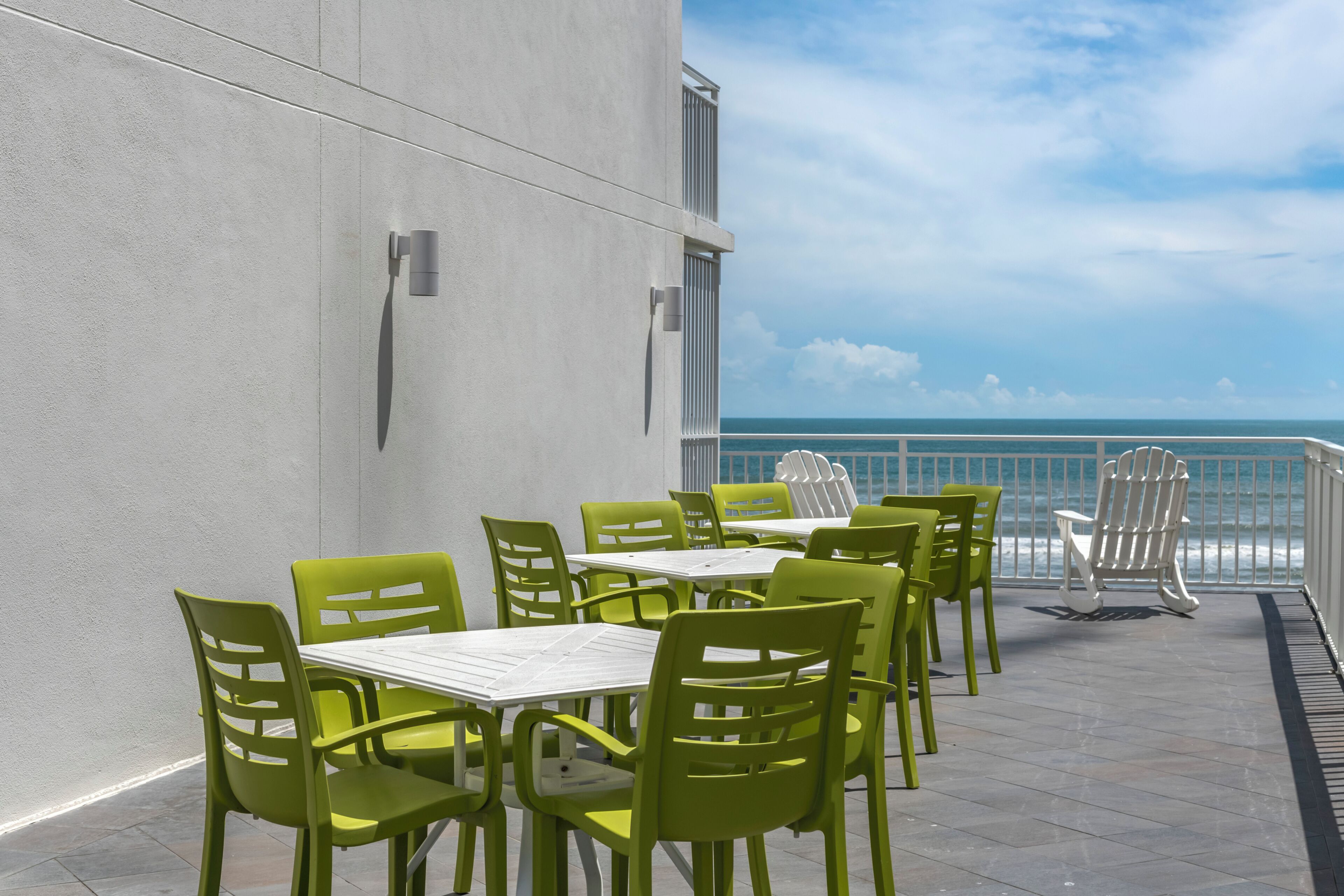 A terrace with green chairs and tables, a white railing, and a view of the ocean.