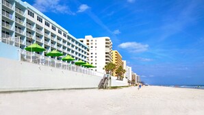 Un hotel frente a la playa con varios balcones, una playa de arena y un cielo azul despejado.