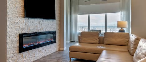 A modern living room with a stone wall, a flat-screen TV, a fireplace, a leather sofa, and a view of the beach through large windows.
