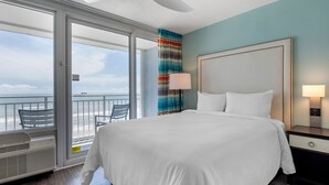 Habitación de hotel con una cama grande, una mesita de noche, una lámpara y vistas al océano a través de una puerta corrediza.
