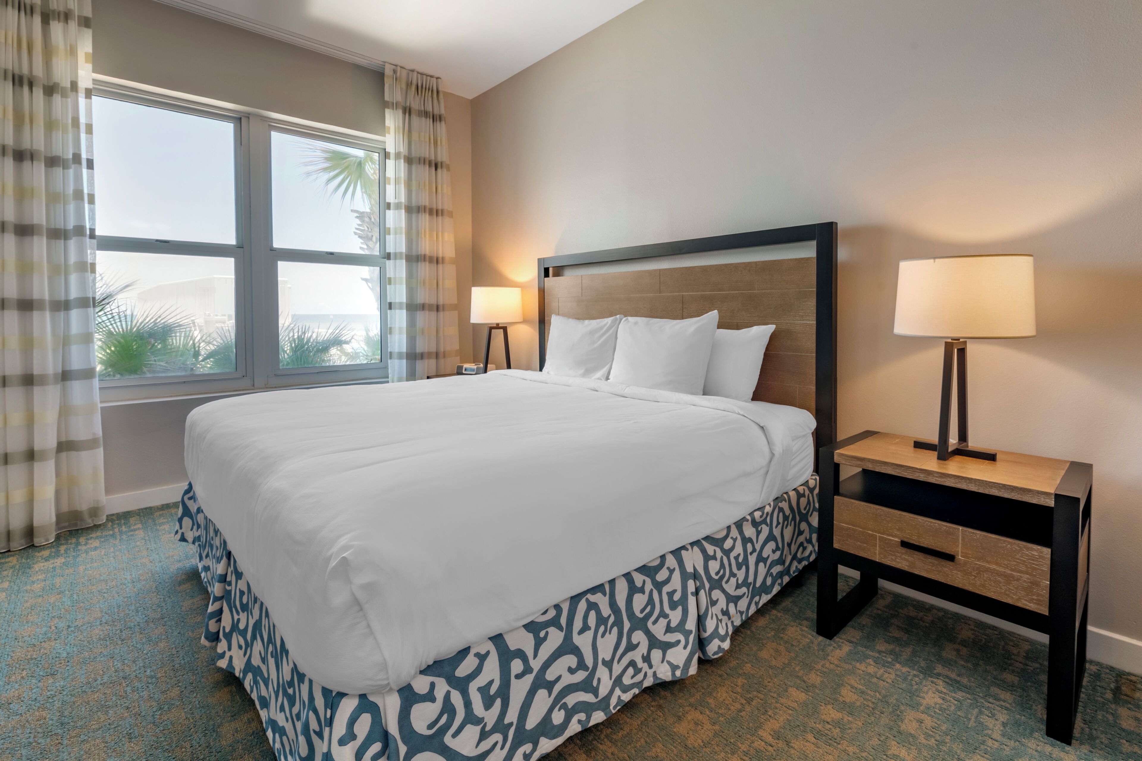 A hotel room with a large bed, two bedside tables with lamps, a window with curtains, and a view of palm trees.