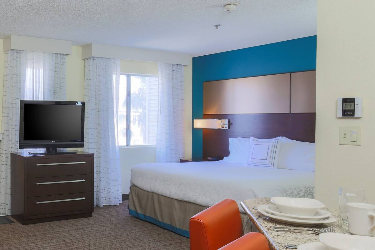 A hotel room with a bed, a TV on a dresser, a dining table with plates and cups, and curtains on the windows.