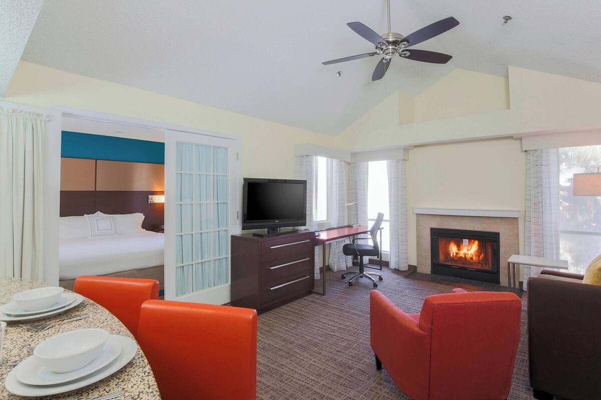 A hotel room with a bed, a desk with a TV, a fireplace, and a dining area with chairs and plates.