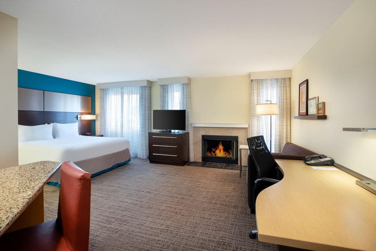 A hotel room with a bed, desk, chair, TV, fireplace, and a small table with a lamp.