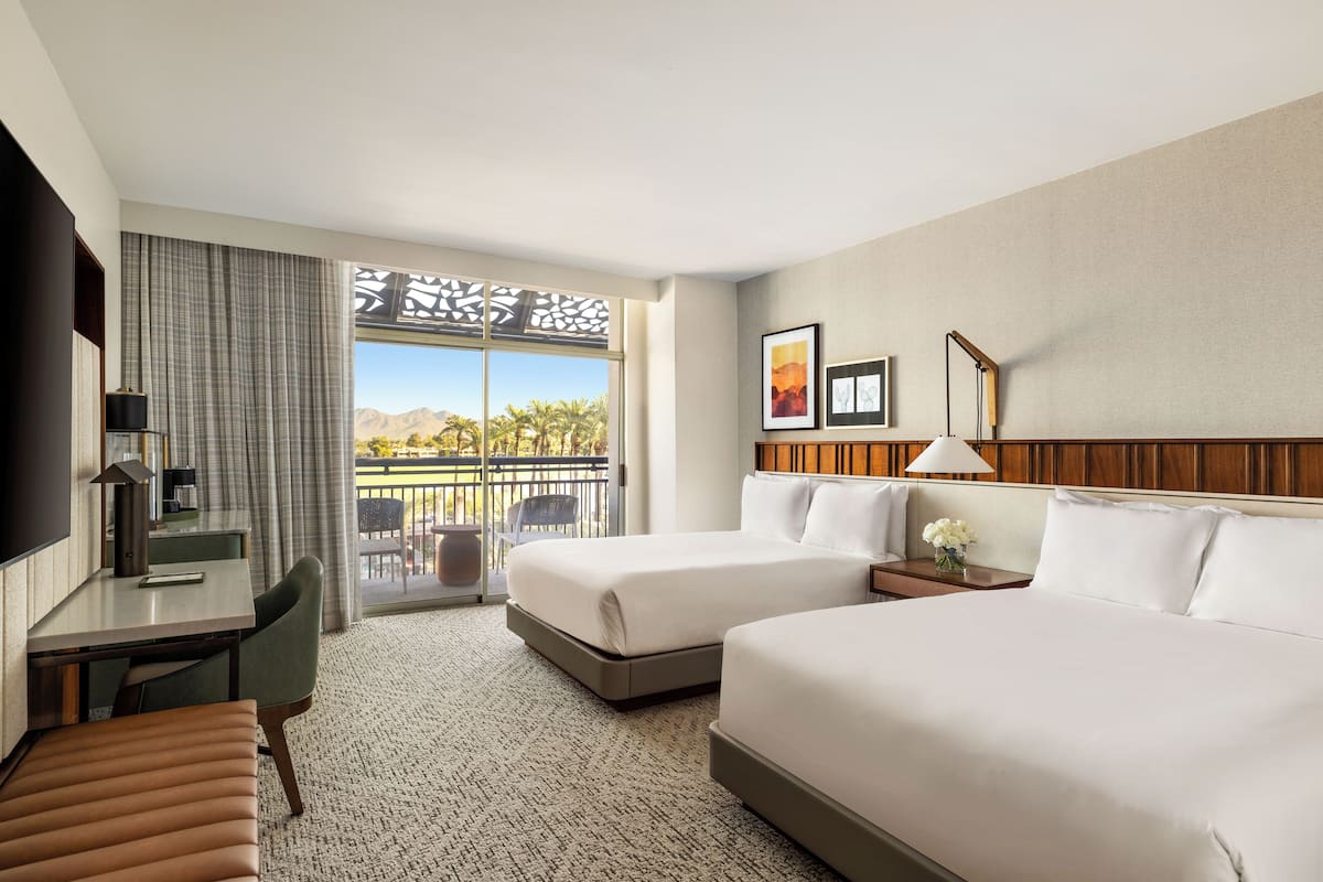 A hotel room with two beds, a desk, a chair, a TV, and a balcony with a view of palm trees and mountains.
