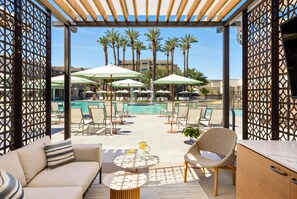A poolside area with lounge chairs, umbrellas, and a view of a building with palm trees.