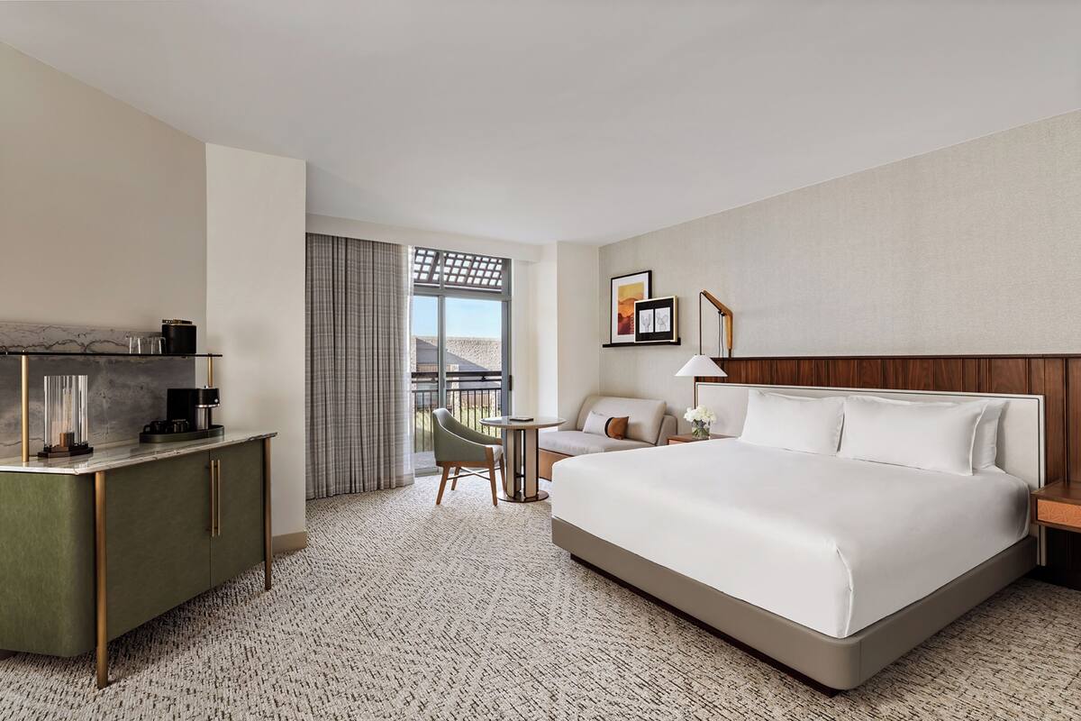 A hotel room with a large bed, a desk with a coffee maker, a chair, a small table, and a balcony with a view.