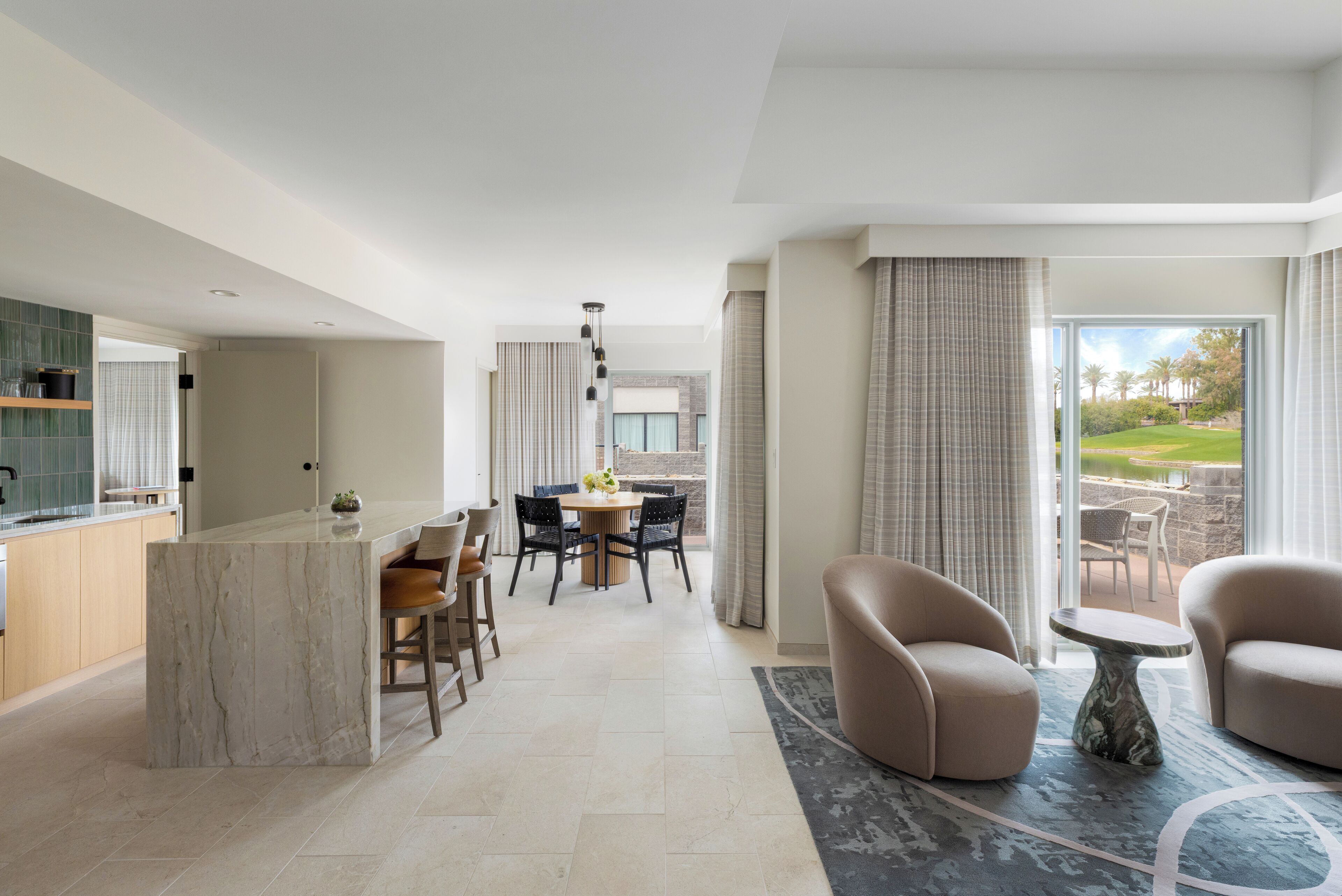 Habitación de hotel moderna con una isla de mármol, zona de comedor y asientos junto a un ventanal con vistas a un jardín ajardinado.