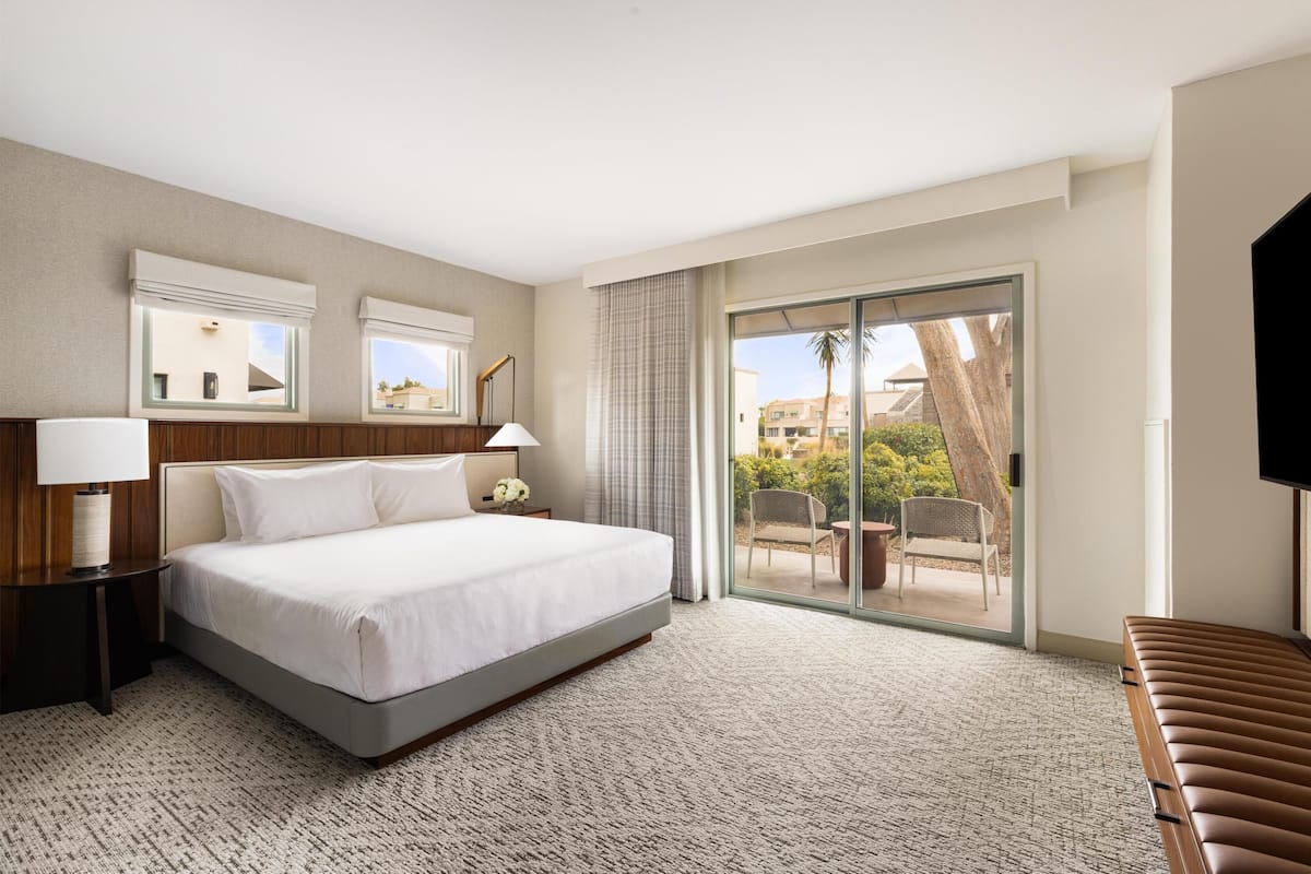 A hotel room with a large bed, a nightstand with a lamp, a TV, a sliding glass door to a balcony, and a view of buildings and trees.