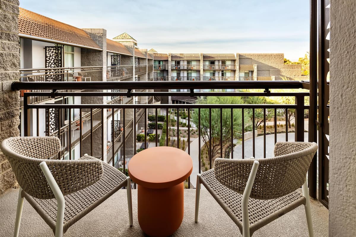 A balcony with wicker chairs and a round table, overlooking a courtyard with buildings and greenery.