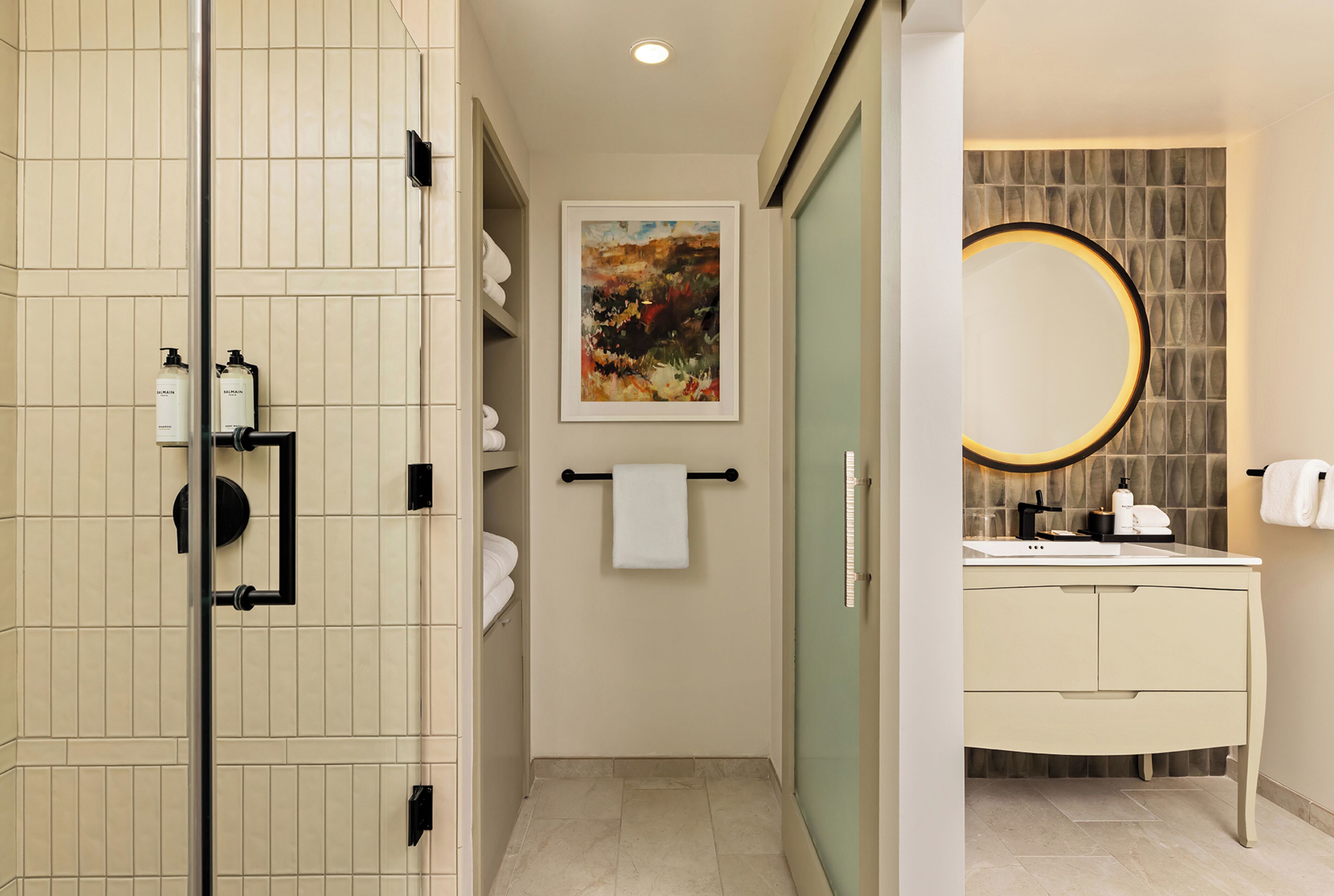 Une salle de bain avec une douche à parois de verre, un porte-serviettes, une œuvre d’art encadrée, un miroir rond et un meuble-lavabo blanc avec des articles de toilette.