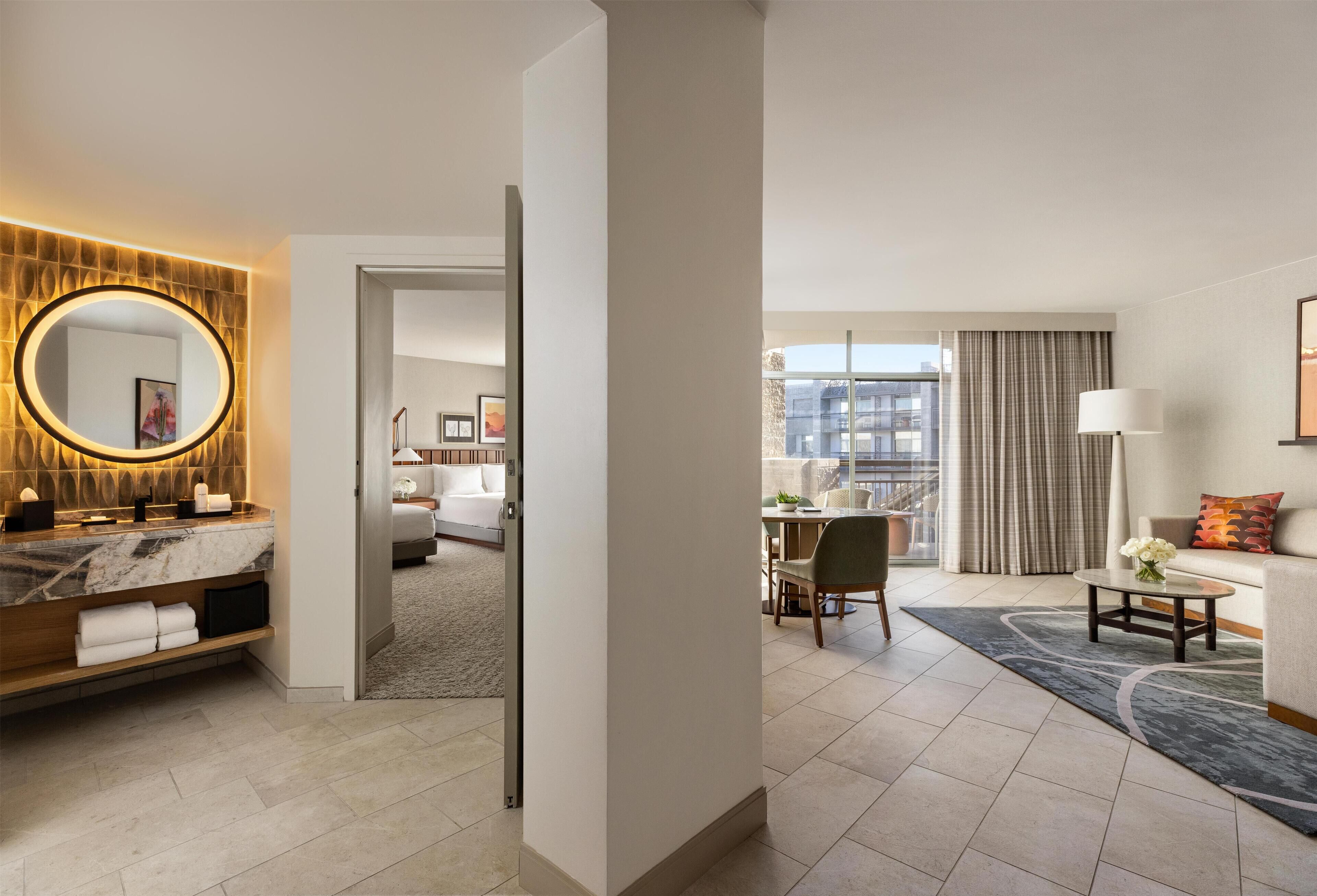 Habitación de hotel con tocador de mármol, espejo redondo, cama, zona de comedor y vistas a la ciudad.