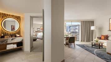 Habitación de hotel con tocador de mármol, espejo redondo, cama, zona de comedor y vistas a la ciudad.
