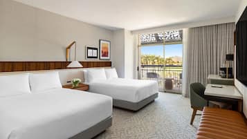 A hotel room with two beds, a desk, a chair, a TV, and a balcony with a view of palm trees and mountains.