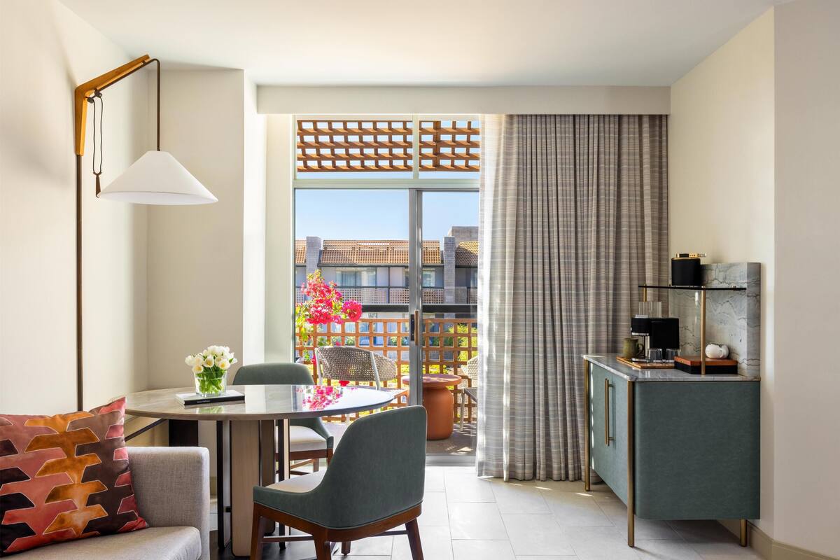 A modern living room with a dining area, a sofa, a table with flowers, a balcony with a view, and a kitchen area with a cabinet.