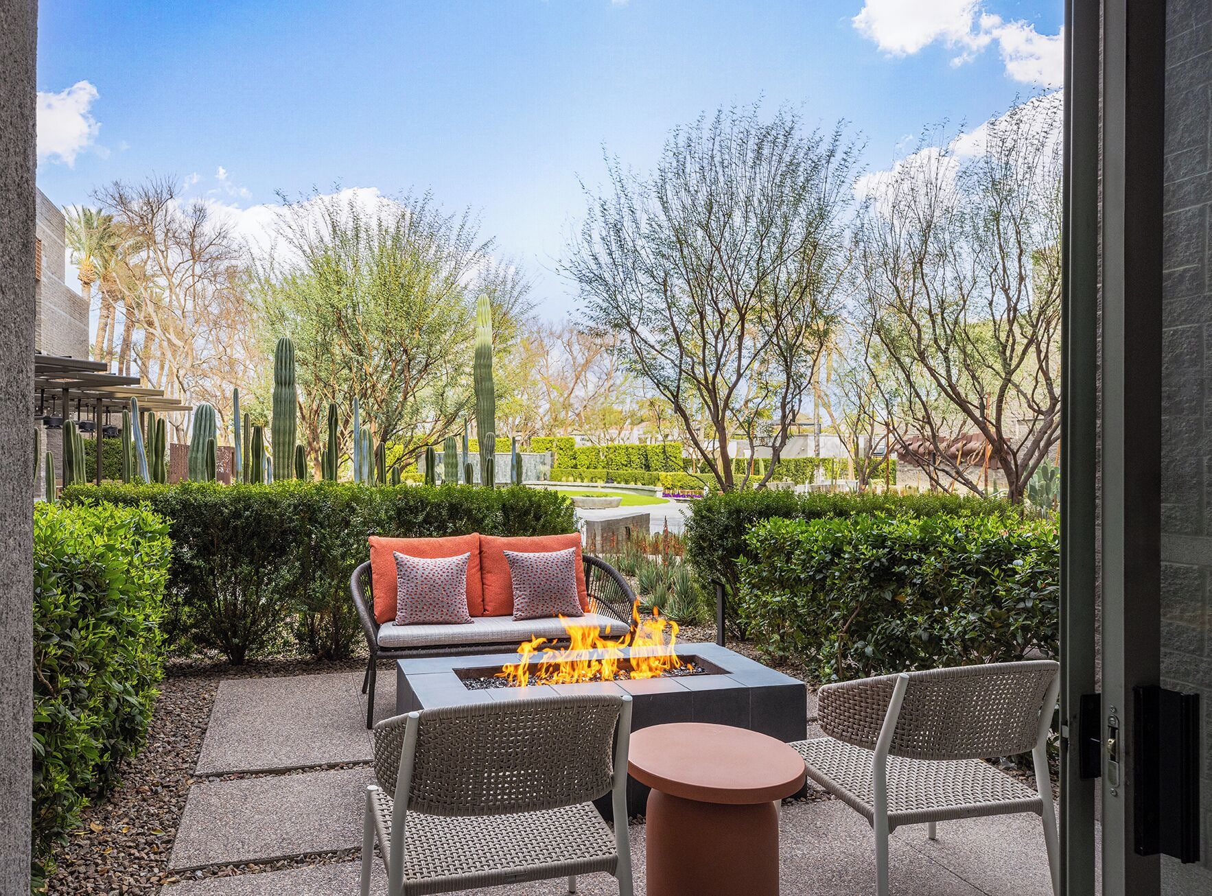 An outdoor patio with a fire pit, wicker furniture, and a view of a landscaped garden with cacti and trees.