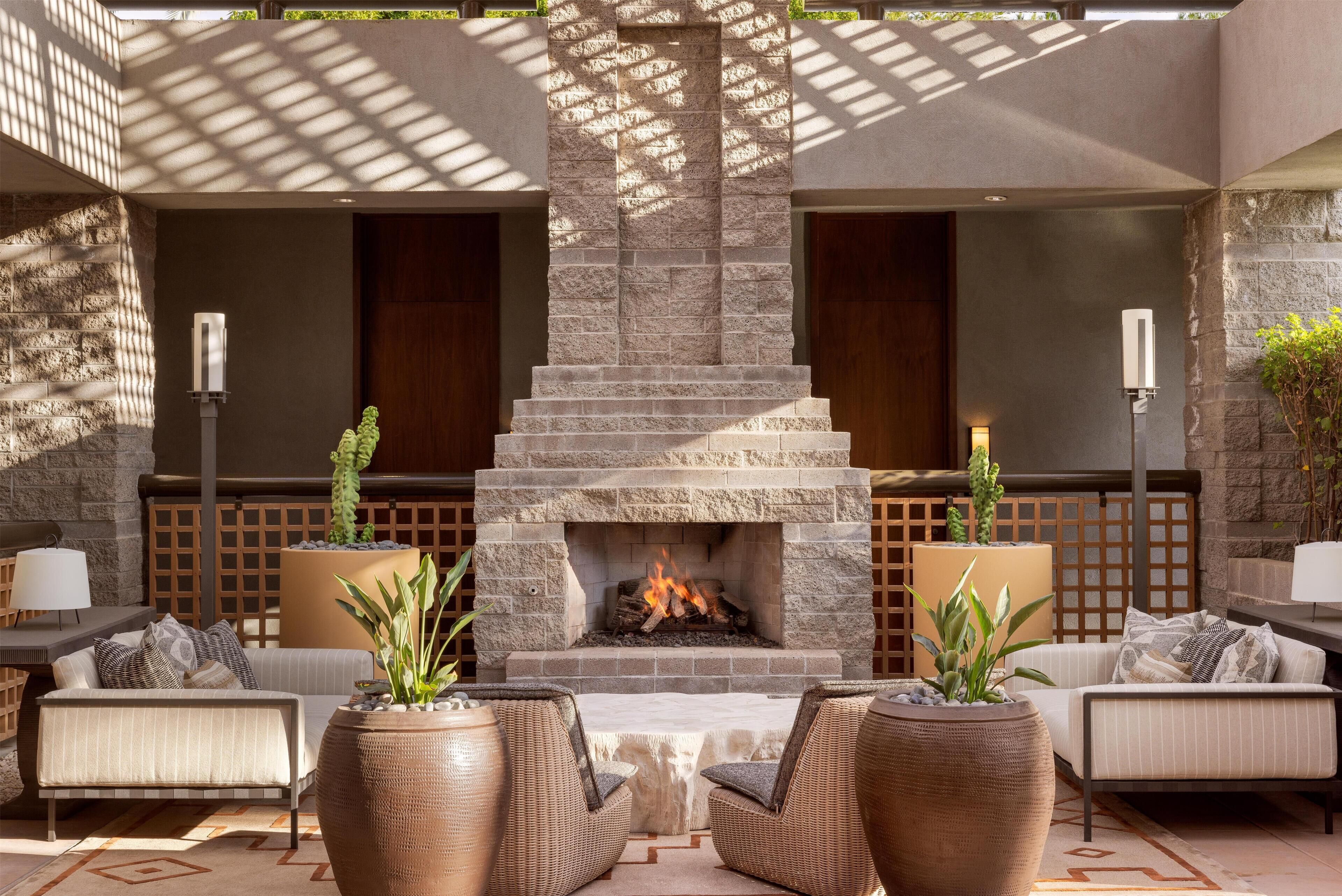 A stone fireplace with a fire, surrounded by wicker furniture and plants, in a sunlit outdoor seating area.