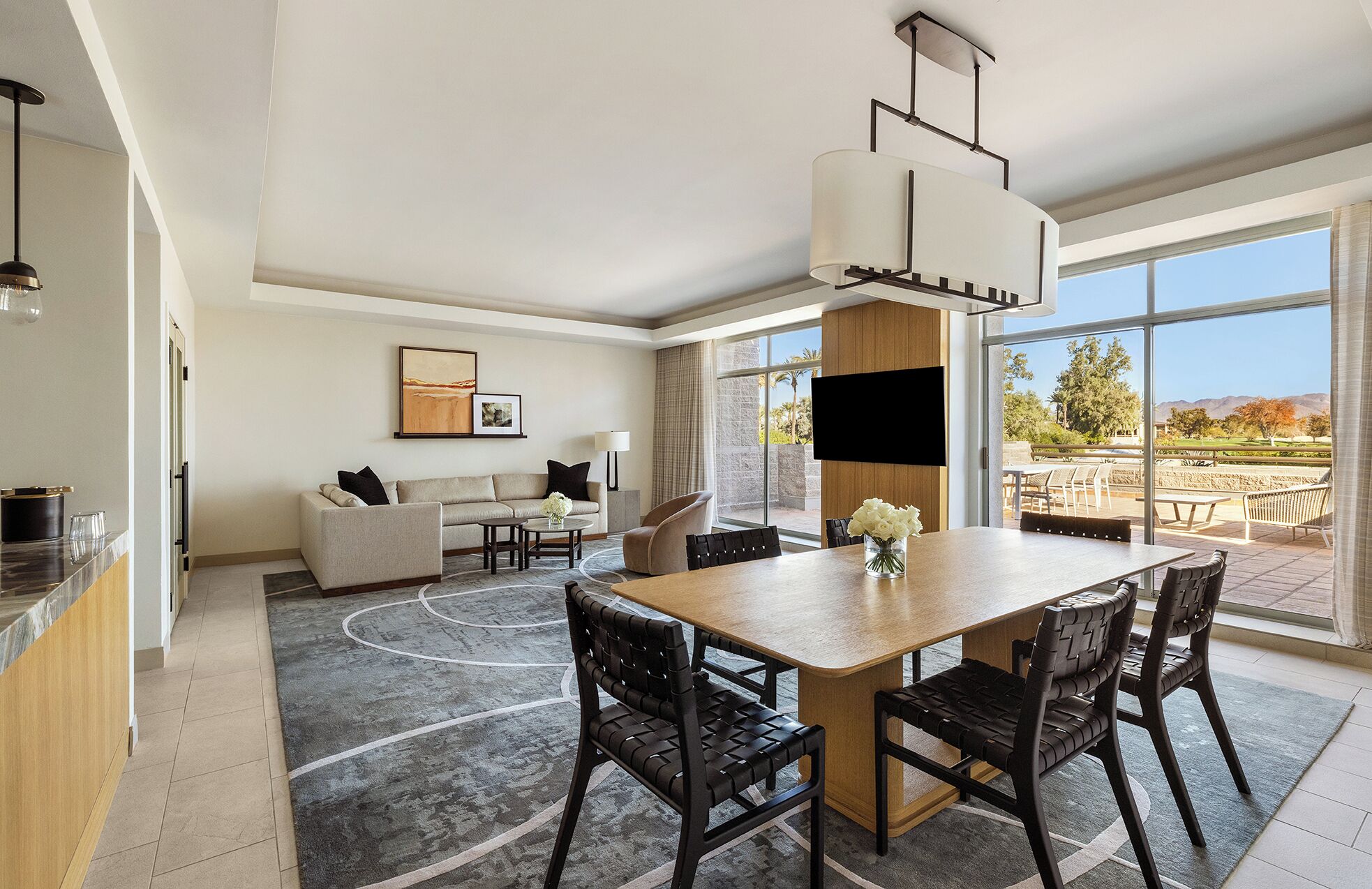 A modern hotel room with a dining area, a sofa set, a flat-screen TV, and a large window offering a view of the outdoors.