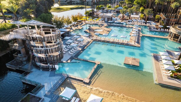 Ein großer, mehrstufiger Pool mit verschiedenen Bereichen und Liegezonen, umgeben von Palmen und einem Golfplatz im Hintergrund.