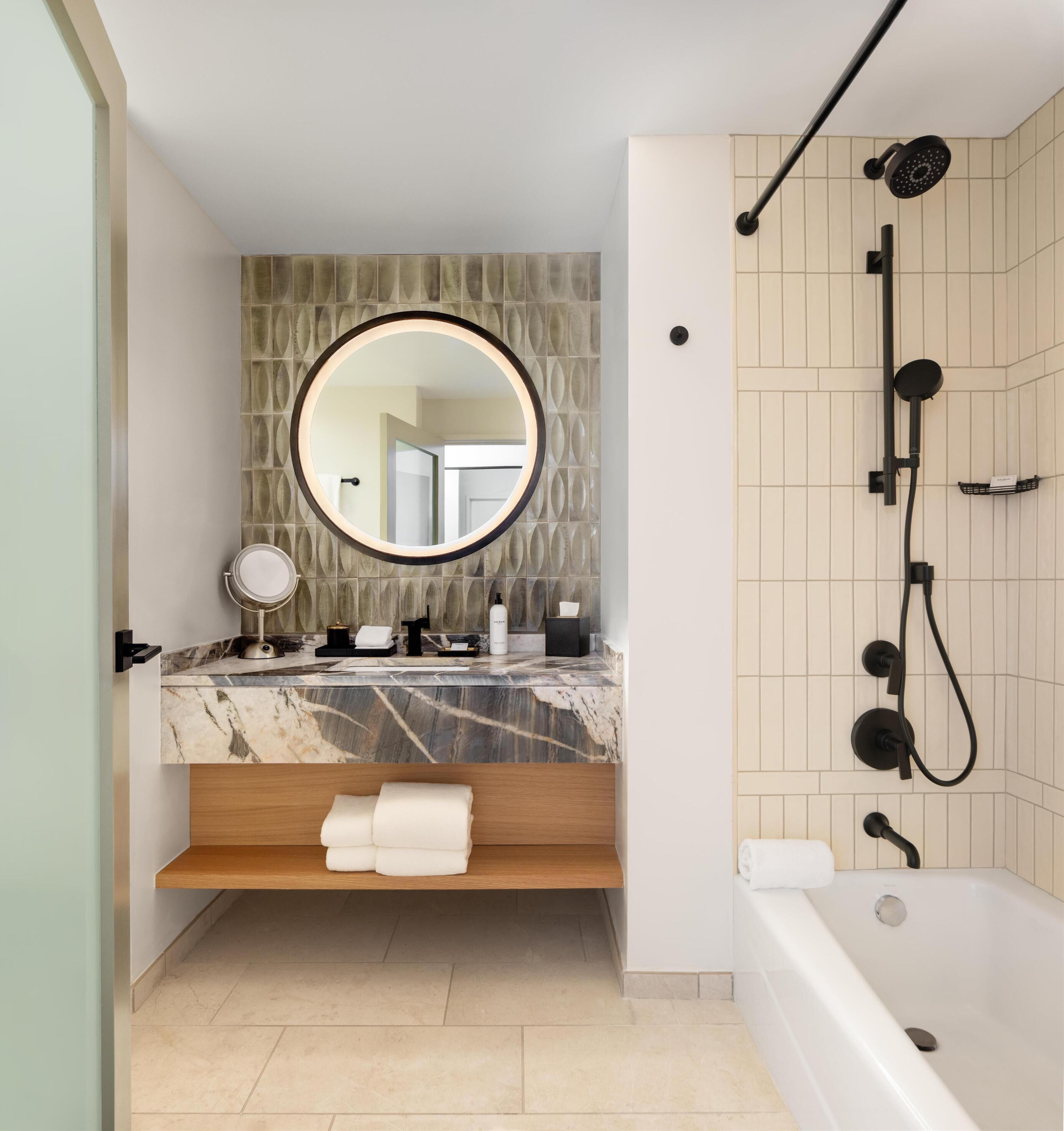 Un baño moderno con tocador de mármol, espejo redondo, estante empotrado, bañera y ducha con múltiples cabezales.