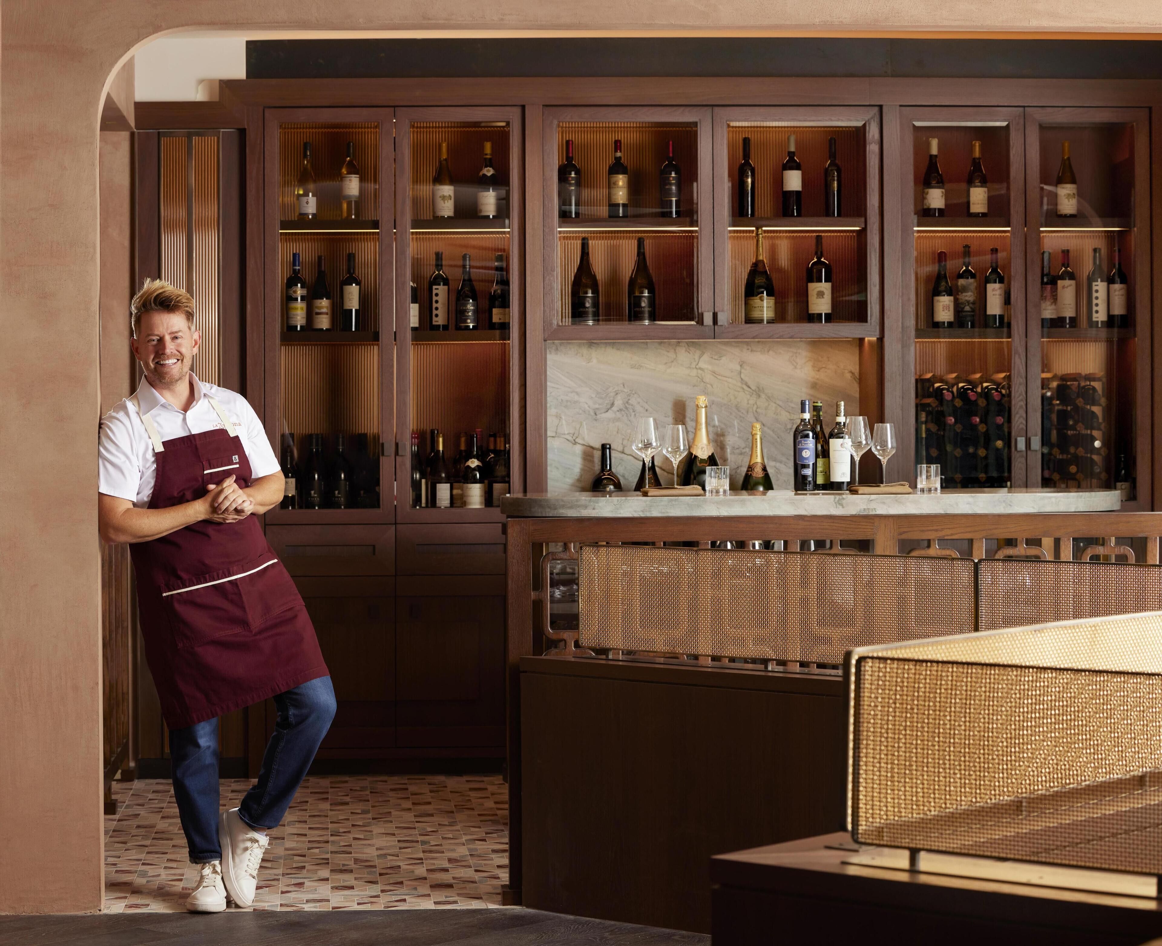 A bar area with shelves of wine bottles, a bar counter, and a person standing with arms crossed.