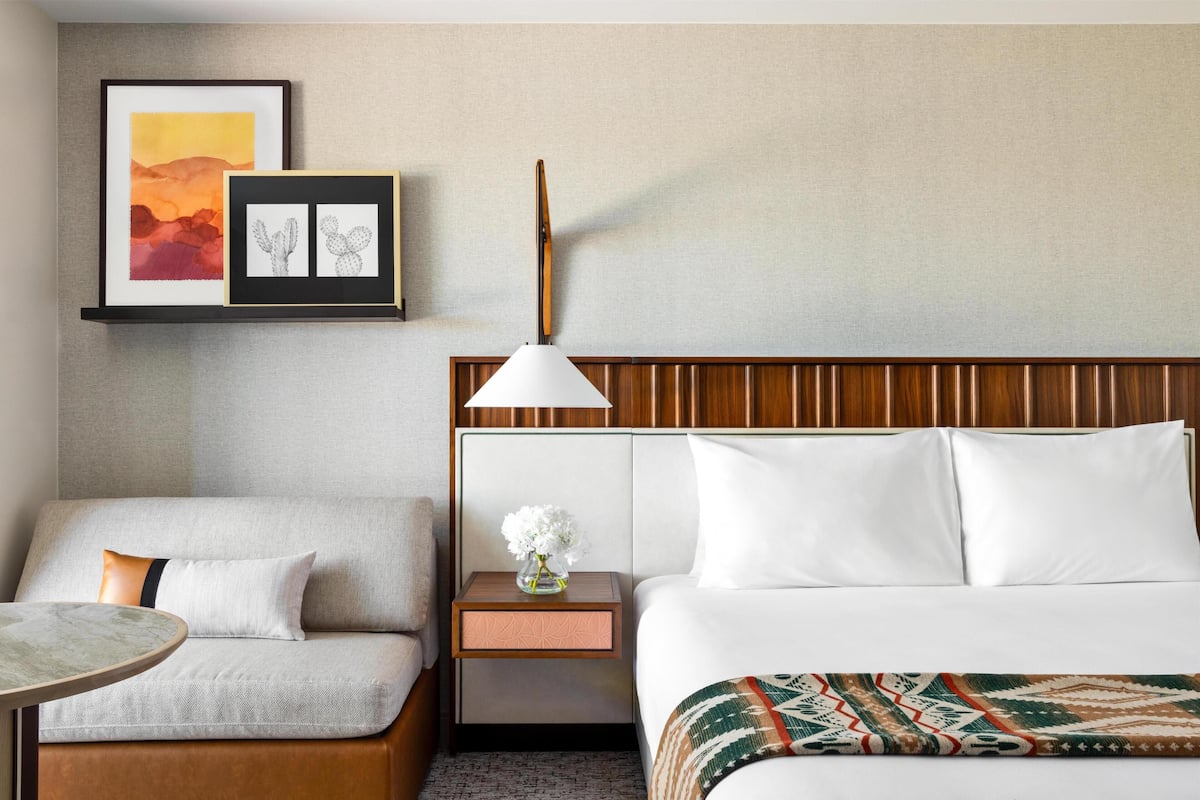 A hotel room with a bed, a sofa, a nightstand with a lamp, and framed artwork on the wall.
