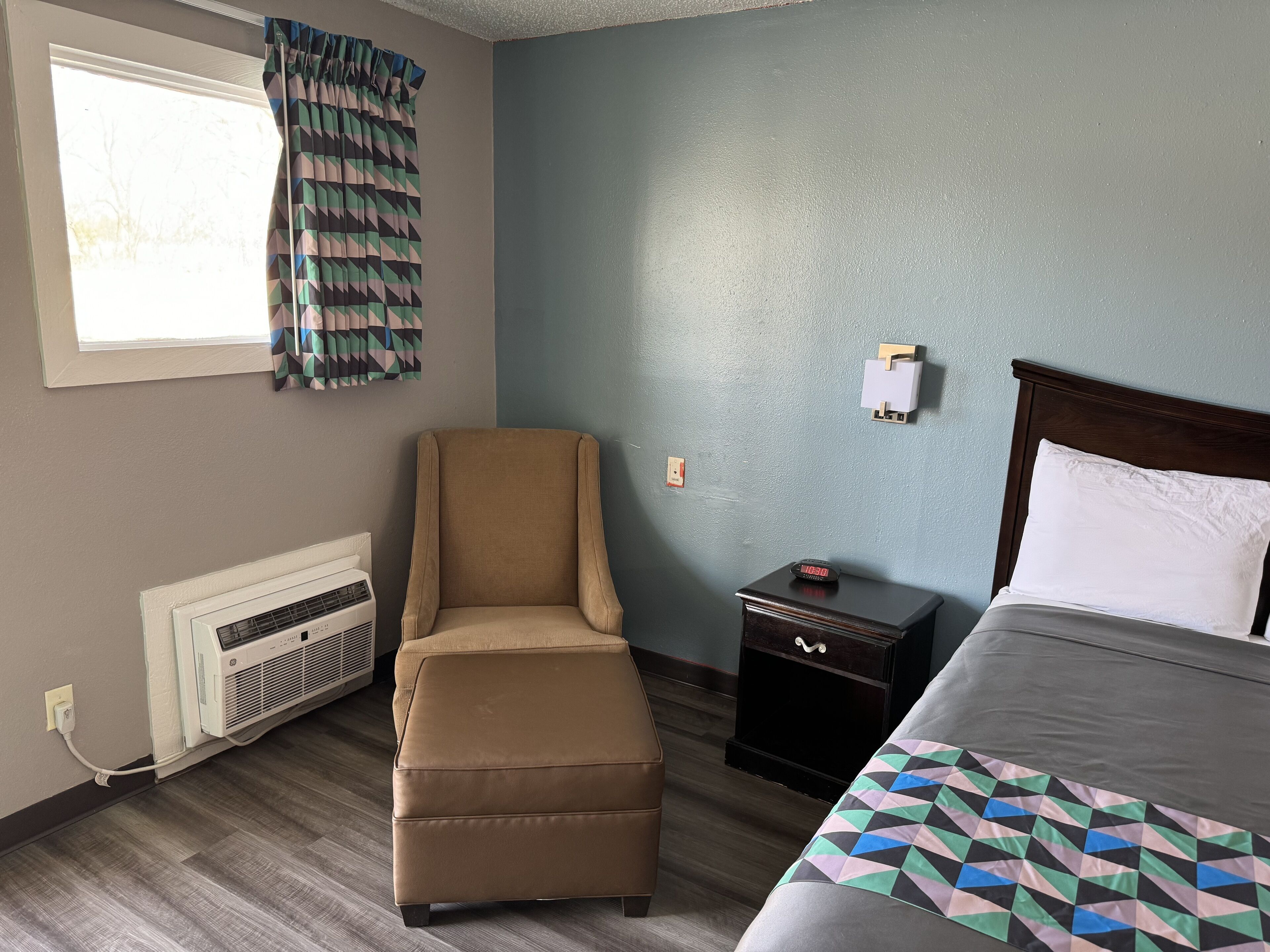 A hotel room with a bed, a chair, a nightstand, a heater, and a window with curtains.