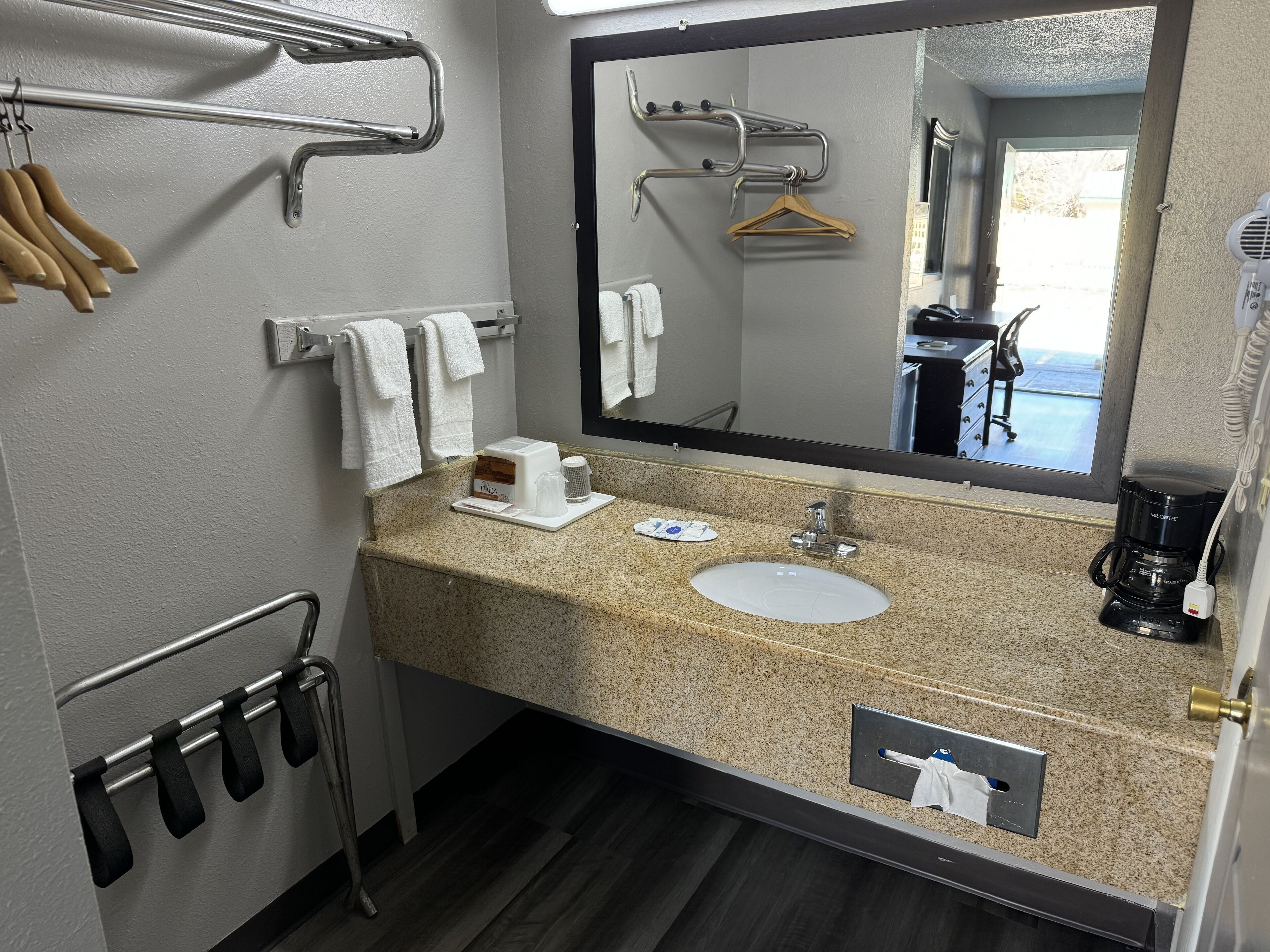 A hotel bathroom with a granite countertop sink, a mirror, a towel rack, and a coffee maker.