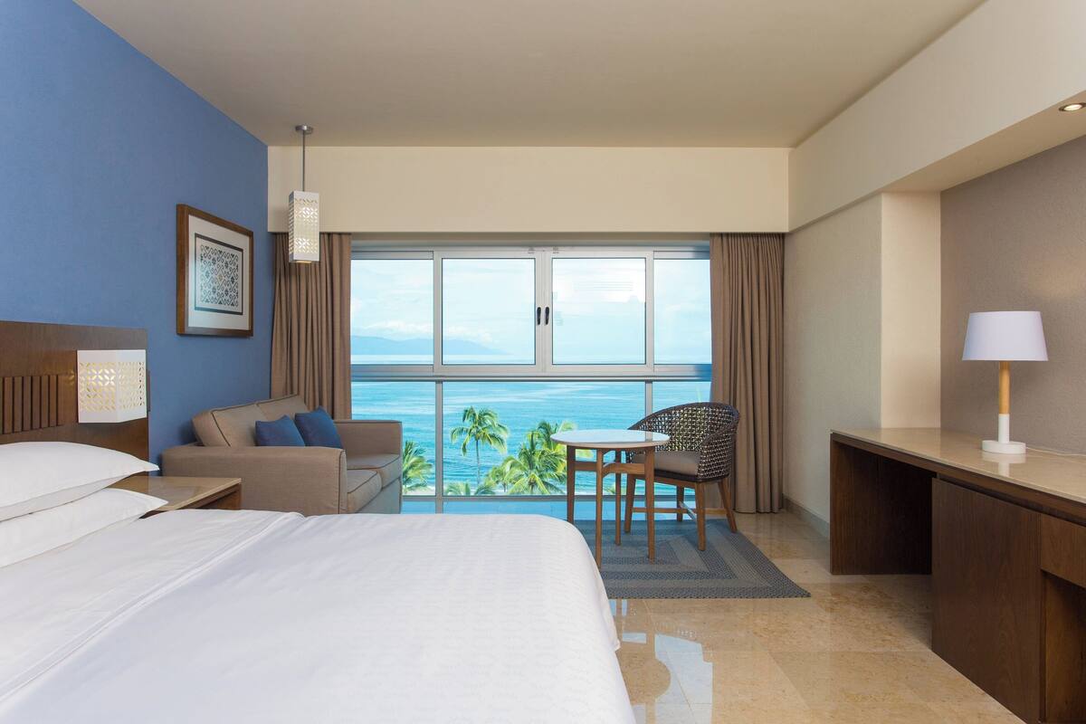 Ein Hotelzimmer mit einem Bett, einem Schreibtisch, einem Sessel, einem kleinen Tisch und Blick auf den Ozean.