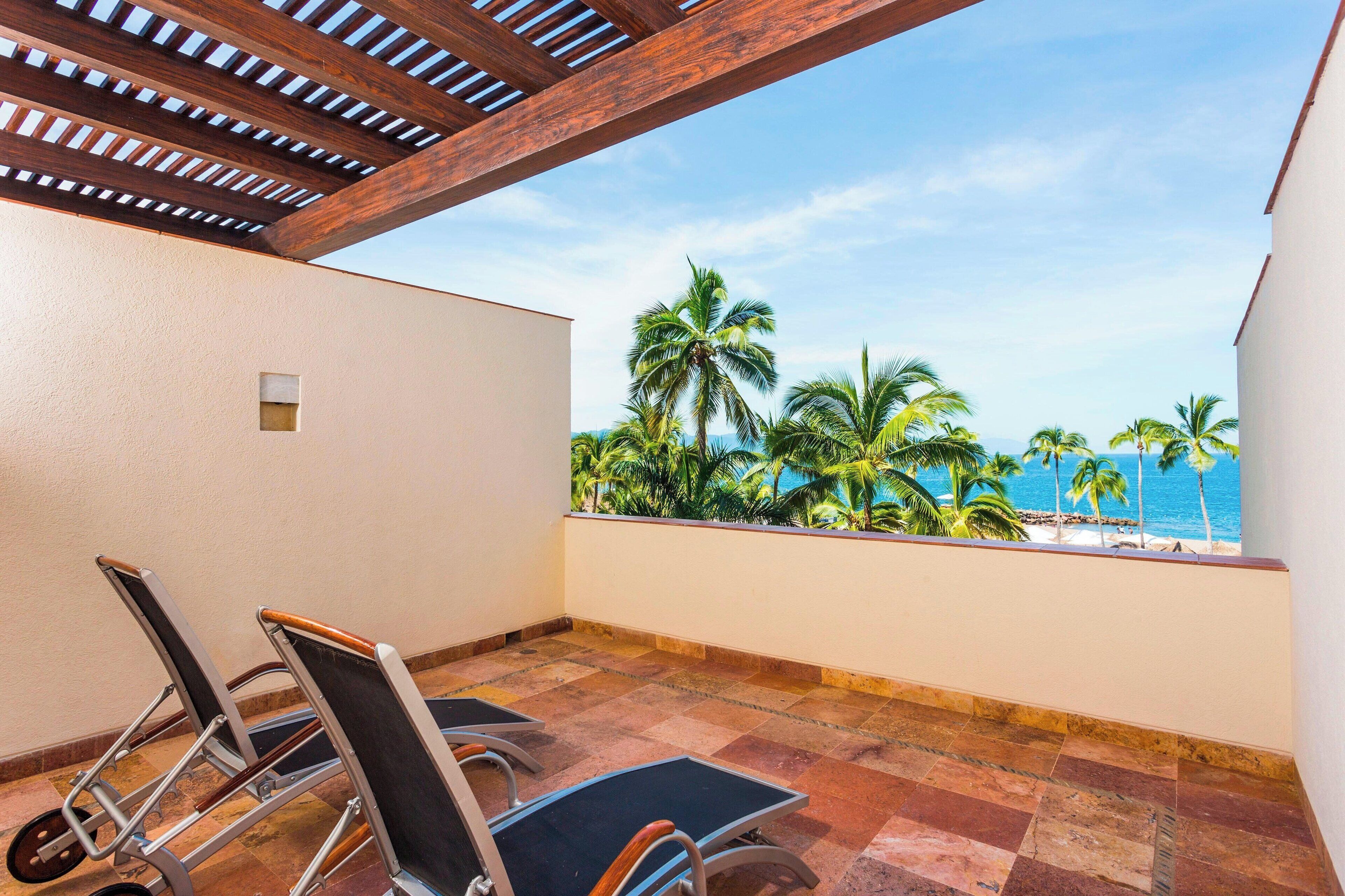 A balcony with two chairs, a view of palm trees and the ocean.