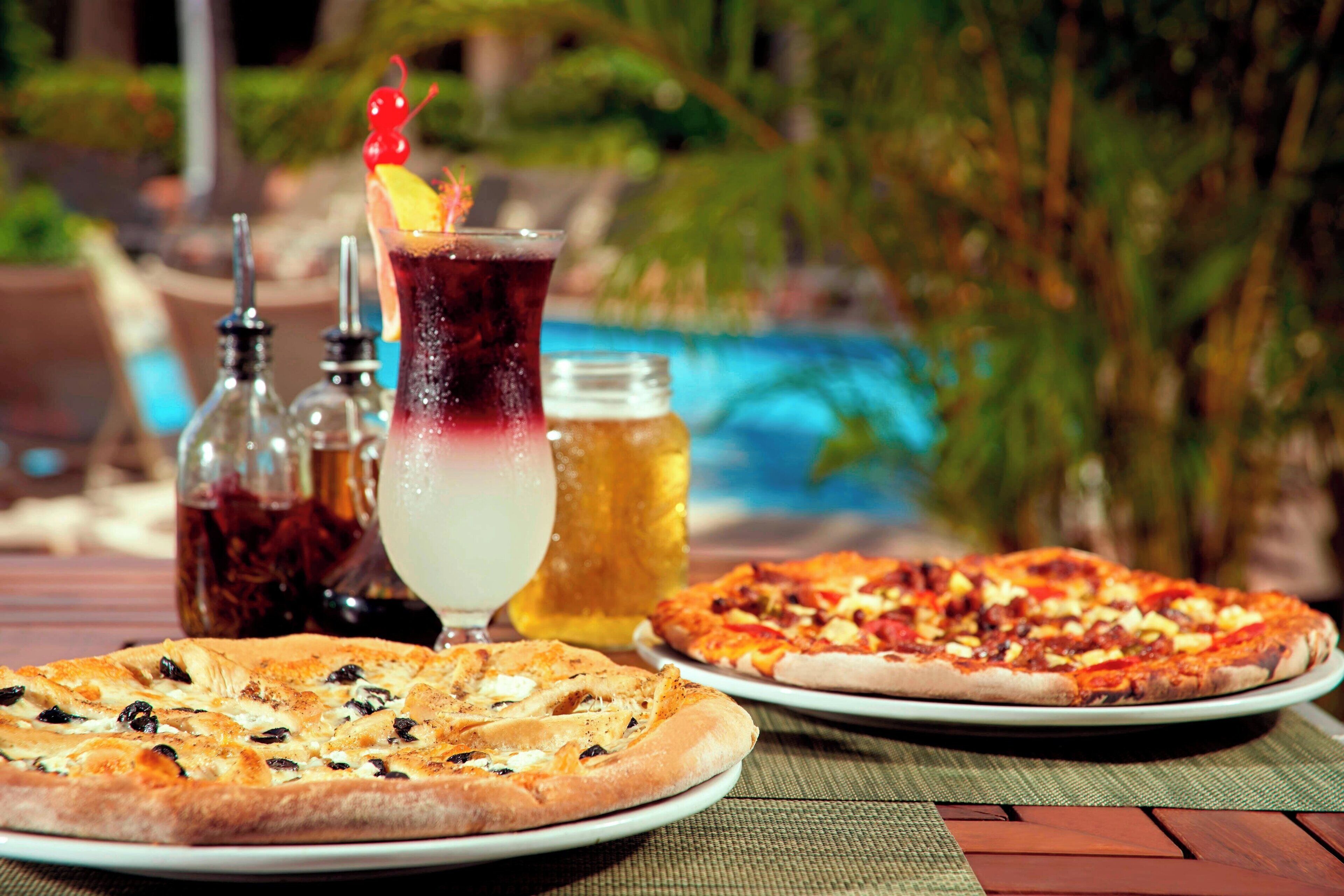 Dos pizzas en platos, una bebida y condimentos sobre una mesa cerca de una piscina.
