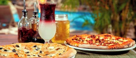 Dos pizzas en platos, una bebida y condimentos sobre una mesa cerca de una piscina.