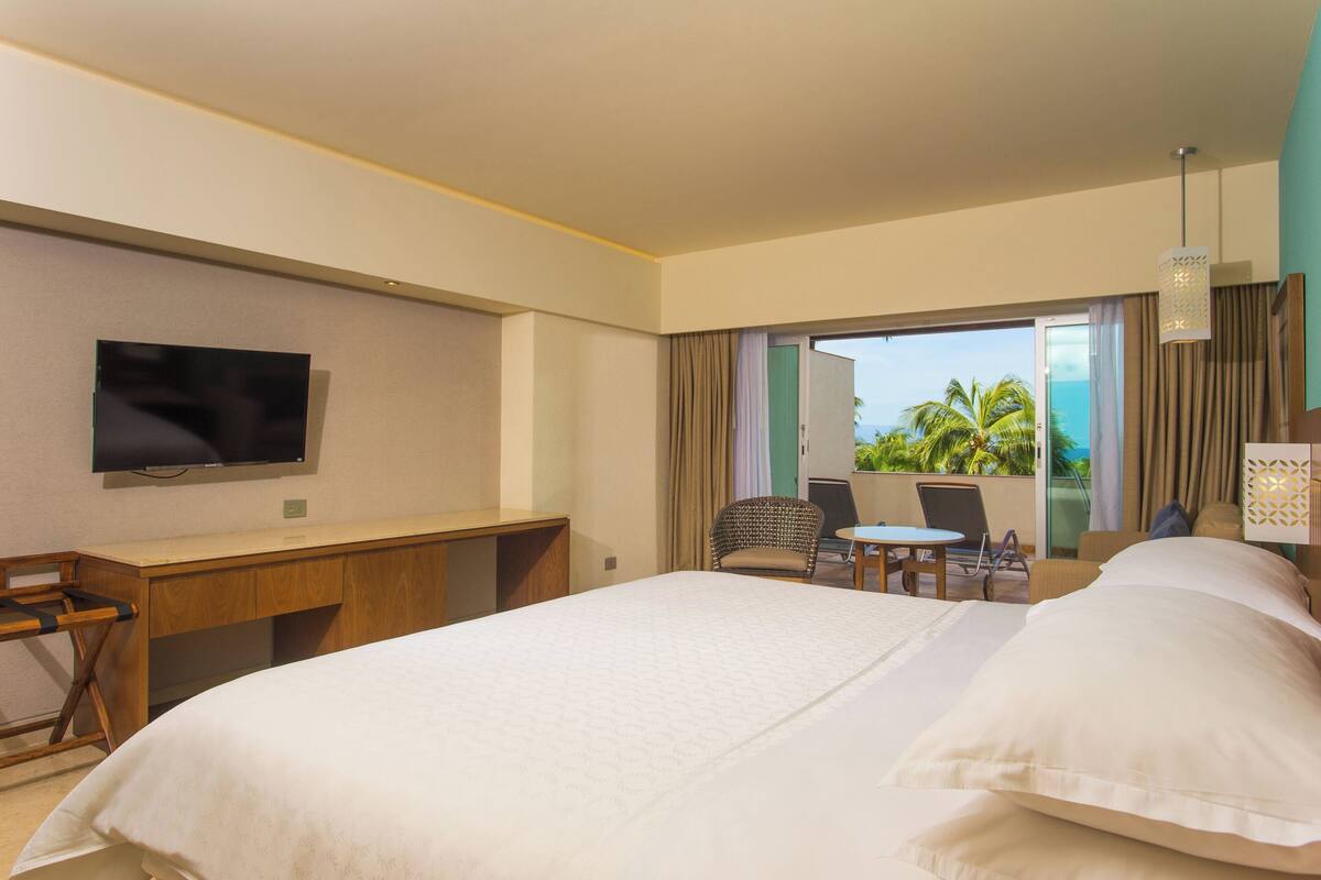 Ein Hotelzimmer mit Bett, Schreibtisch, Fernseher, Sessel, Tisch und Blick ins Freie.