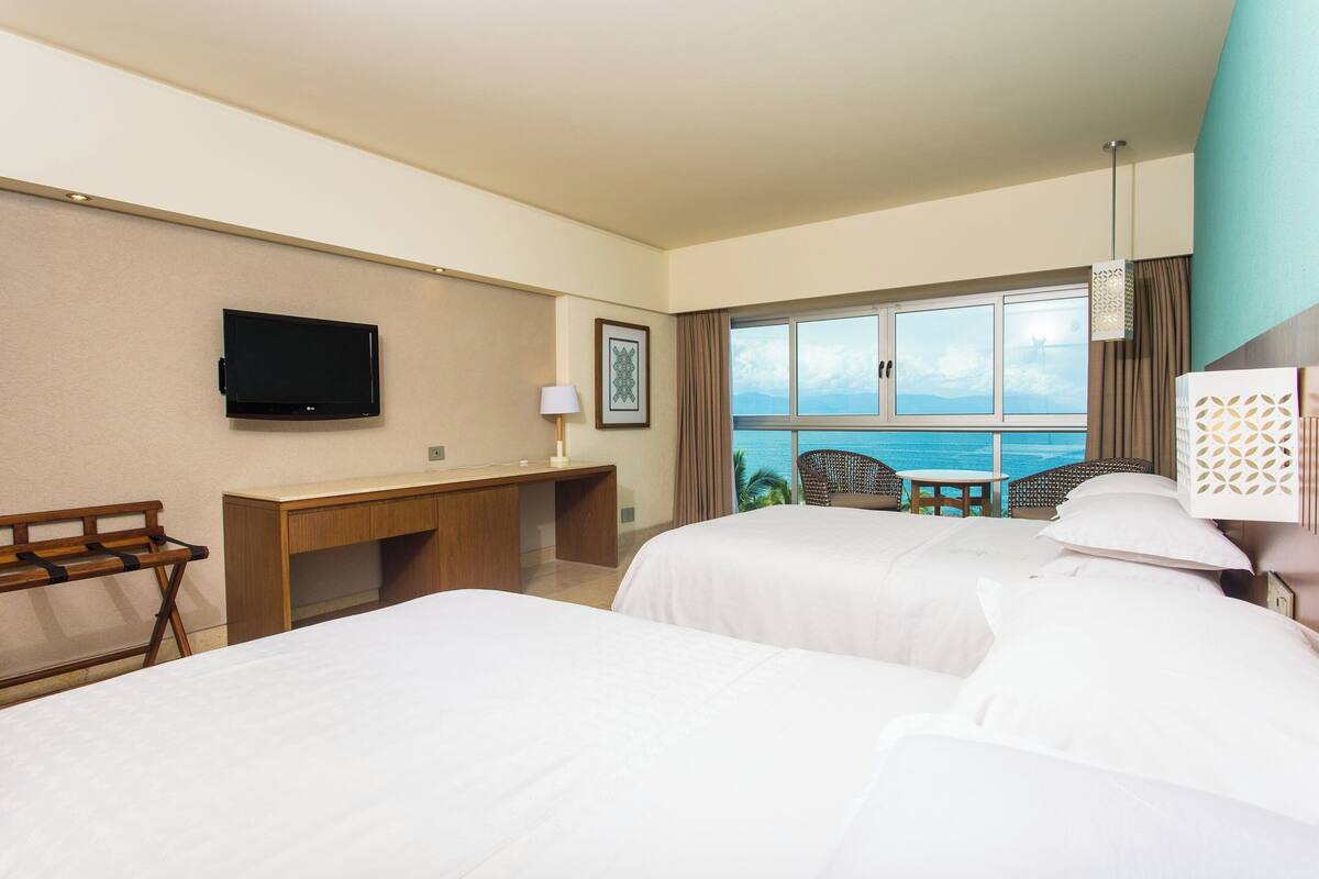 Ein Hotelzimmer mit zwei Betten, einem Schreibtisch, einem Fernseher und einem Balkon mit Meerblick.