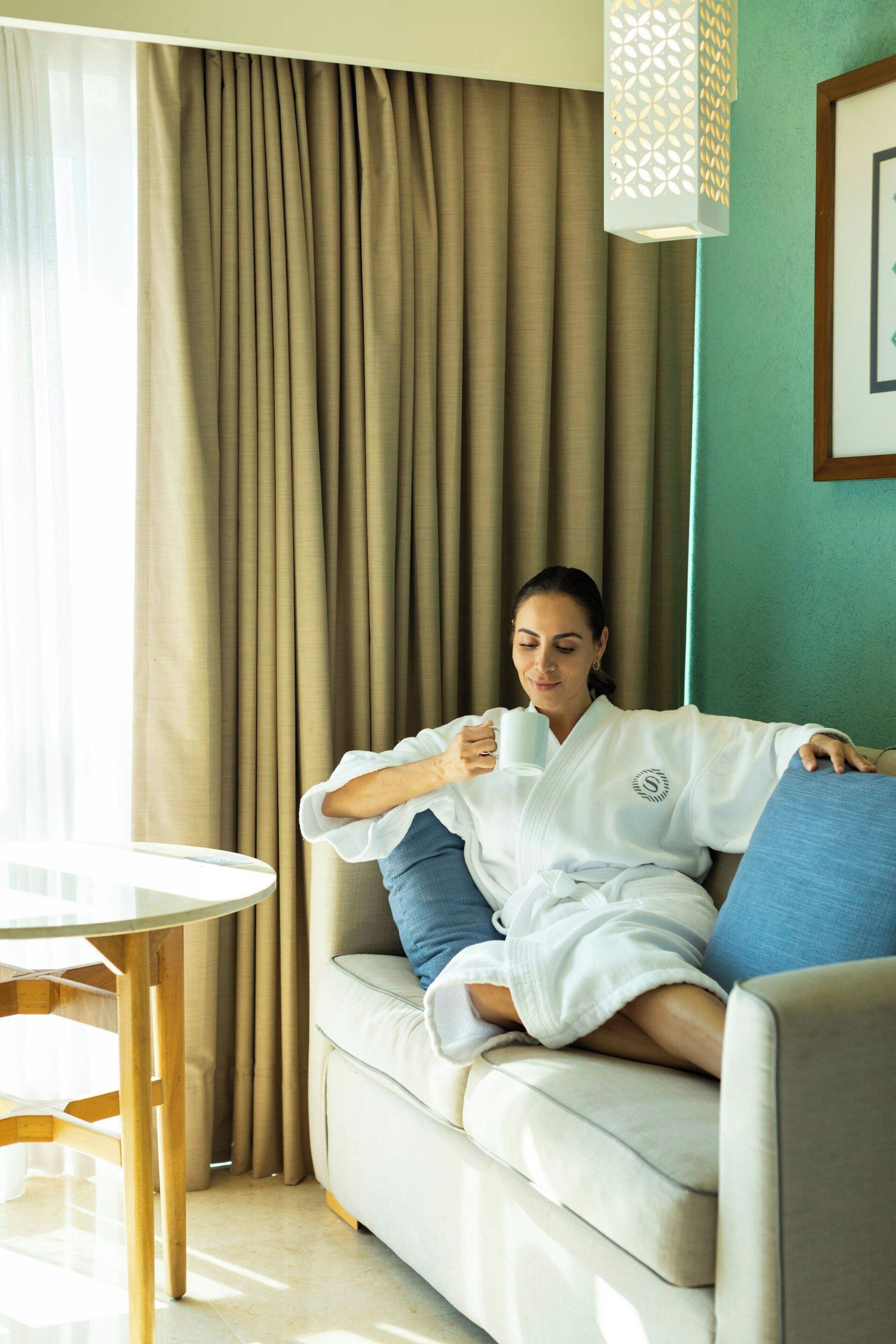 A person in a white robe sitting on a beige sofa with blue cushions, holding a white mug.